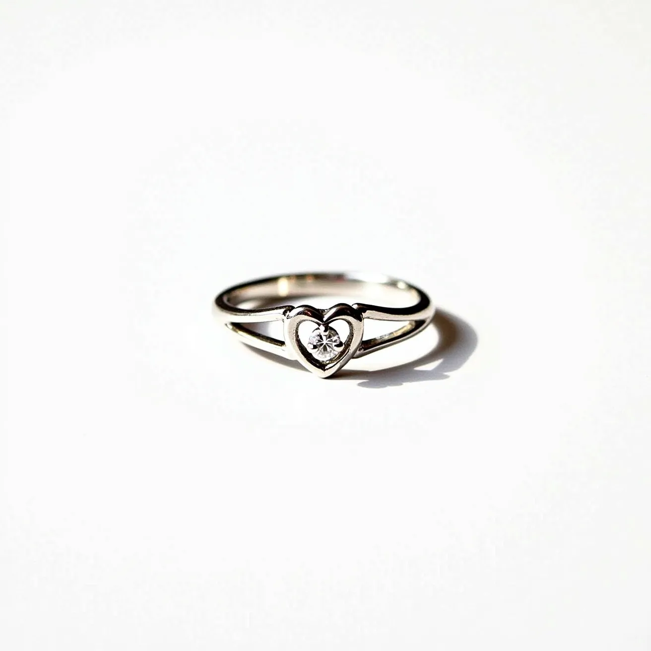 This silver promise ring features a delicate heart-shaped setting, crafted from polished silver that exudes a simple yet elegant charm. At the center of the heart is a round, brilliant-cut gemstone, likely a diamond or cubic zirconia, that sparkles with clarity. The stone is securely held in place, enhancing its brilliance and adding a touch of sophistication to the overall design. The band gracefully splits as it approaches the centerpiece, providing additional emphasis and framing the heart beautifully. This piece exemplifies the understated elegance often desired in promise rings.