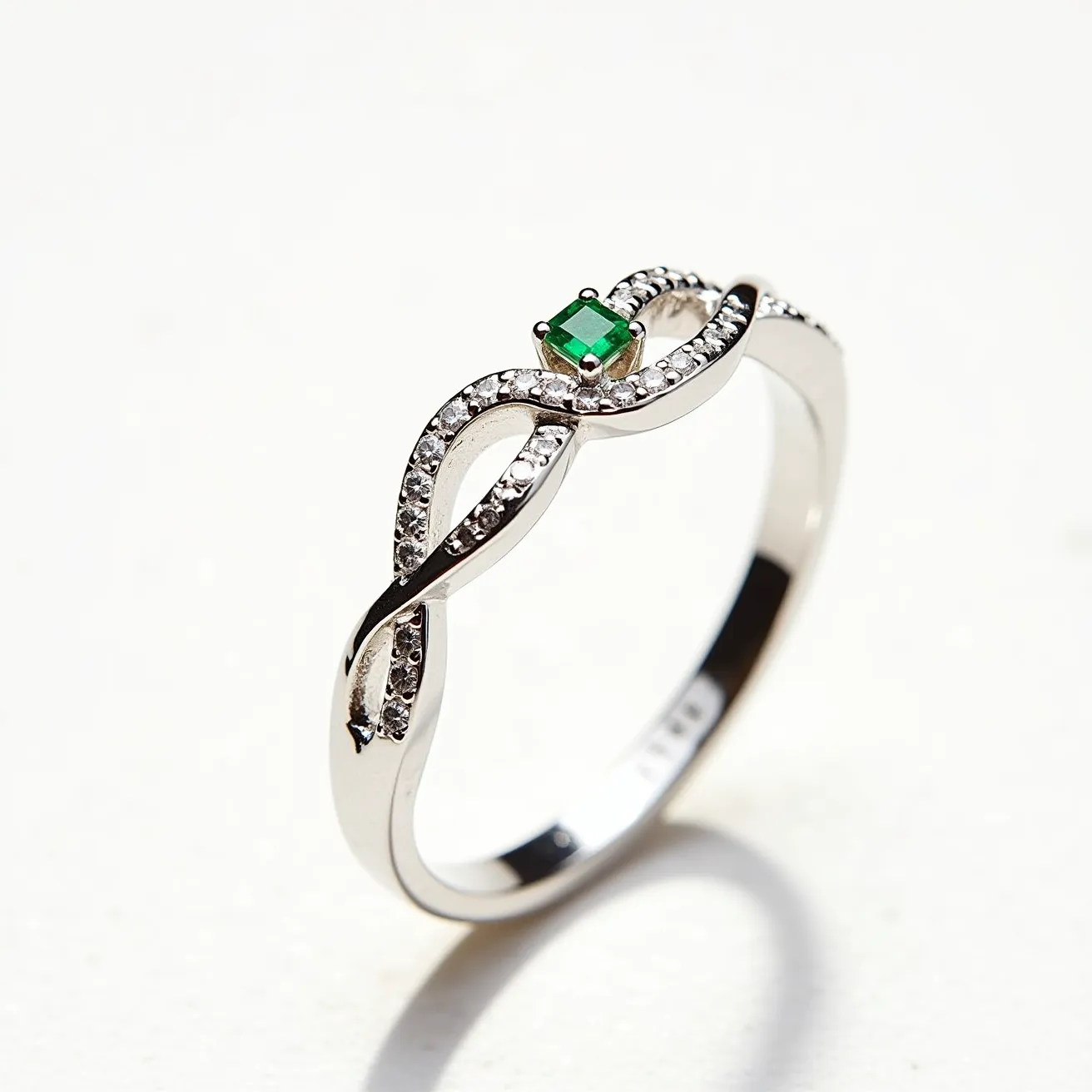 This silver promise ring features a delicate and elegant twisted band design, crafted from polished silver material. Adorning the ring is a small, square-cut green gemstone, likely an emerald, set in a secure prong setting at the center of the twist. The twist is further embellished with small sparkling stones, possibly diamonds or cubic zirconia, enhancing the overall brilliance of the piece. The ring does not feature any clasps or attachments, emphasizing its simplicity and sophistication.