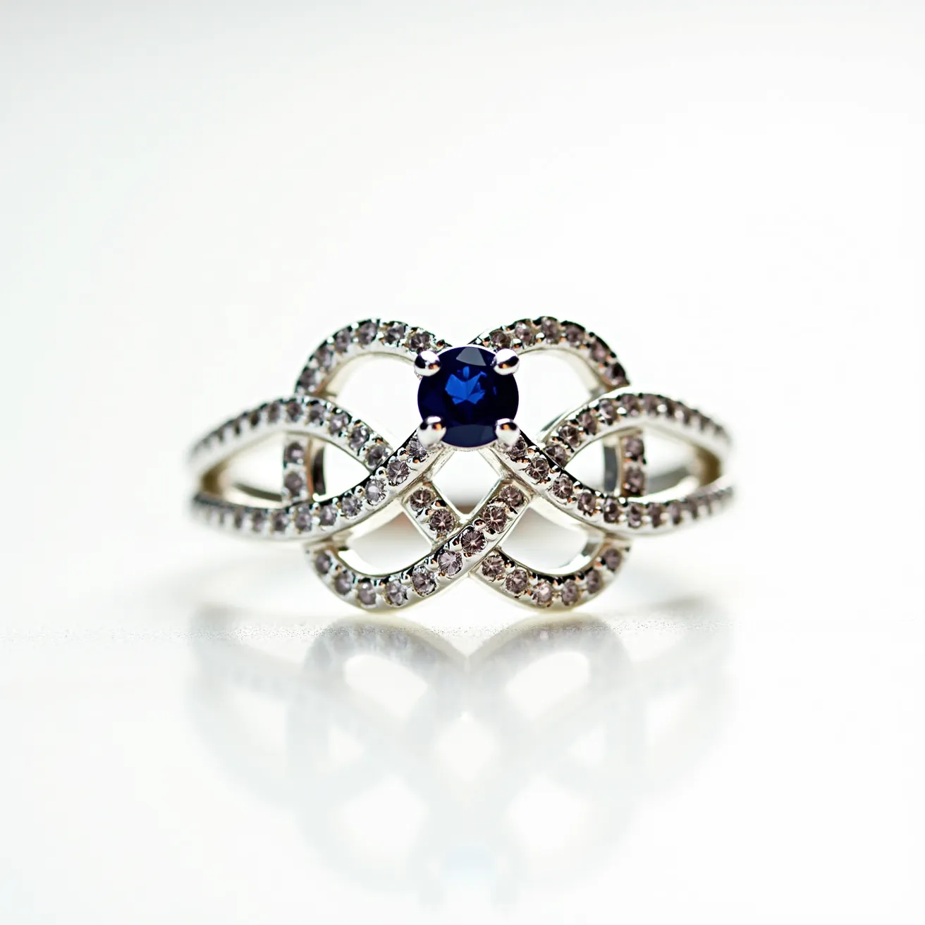 This silver promise ring features an intricate infinity knot design adorned with numerous small, round-cut clear stones, likely cubic zirconias, set along the loops and curves of the band. At the center, a single deep blue round-cut gemstone, possibly a sapphire or sapphire simulant, is securely held in place by a four-prong setting, adding a striking contrast to the overall design. The combination of the silver band and the shining stones creates an elegant and timeless piece, encapsulating a sentiment of commitment and eternity.