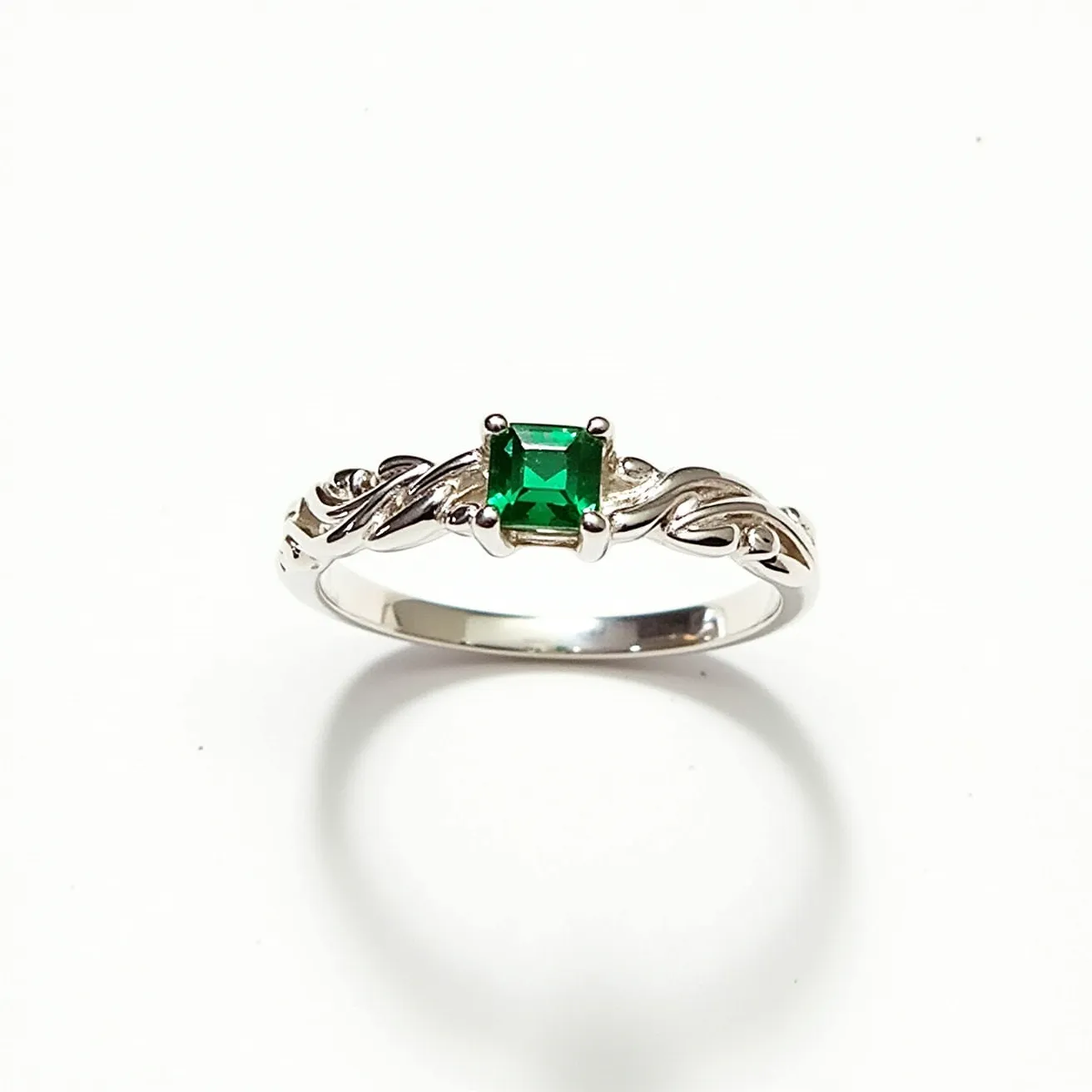 This silver ring features an intricately designed band with a Celtic knot pattern, showcasing a centrally set square-cut green gemstone held securely by a four-prong setting. The green gem, possibly an emerald or a similarly colored stone, adds a vibrant touch to the polished silver band, enhancing its elegance and appeal. The prongs not only hold the gem firmly but also allow light to pass through, highlighting the stone’s brilliance. The detailed craftsmanship of the band complements the gemstone, creating a balanced and attractive piece of jewelry.