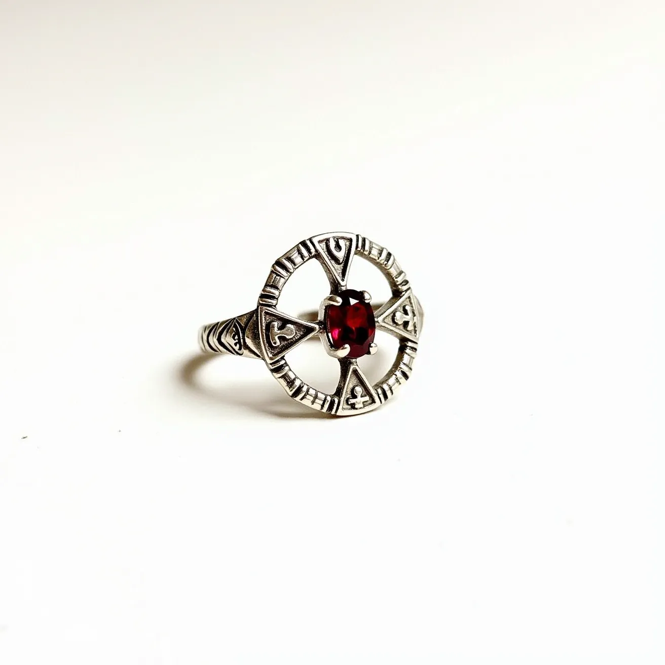 This silver ring showcases intricate Celtic-style motifs around its circular design, highlighted by a vibrant red gemstone at the center. The gemstone appears to be an oval or cushion cut, securely held in place by a four-prong setting. The silver band features decorative engravings that complement the central design, enhancing the overall aesthetic of the piece. There are no additional clasps or attachments on the ring, making it a singularly crafted piece that accentuates the beauty of the materials used.