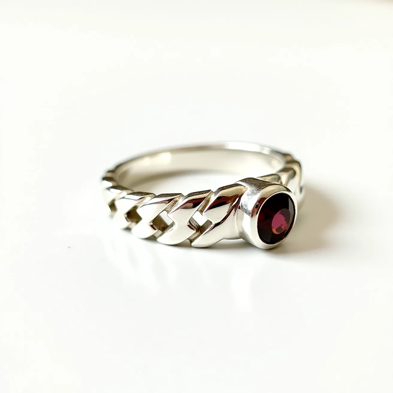 This silver ring features an intricately woven band with a polished finish, showcasing a geometric design that adds texture and depth. At the center of the ring is a round, deep red gemstone, likely a garnet, which is expertly cut to enhance its natural brilliance and is securely set in a bezel setting. The combination of the silver metal and the rich hue of the gemstone creates a striking contrast, making this piece both elegant and visually appealing.