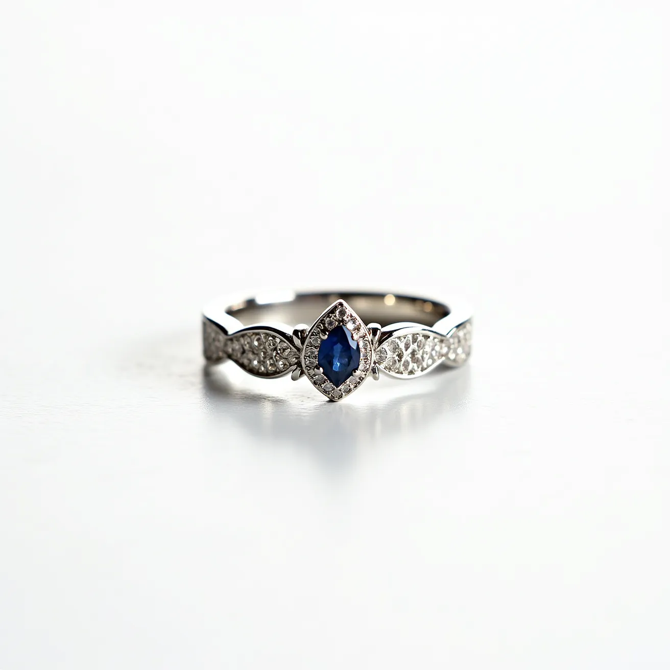 This silver wedding ring features a central blue gemstone, likely a sapphire, cut in an oval shape and set in a prong setting. The band is embellished with small, round-cut diamonds arranged in intricate patterns on both sides of the central stone, providing an elegant contrast. The ring showcases a high-polish finish, adding to its overall brilliance and sophistication. There is no visible clasp or attachment, as it is designed in a continuous circular band typical of wedding rings.