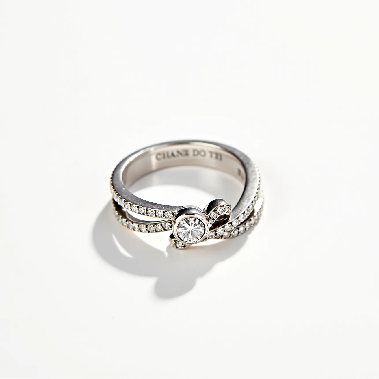 This silver wedding ring features a central round-cut stone set in a bezel setting, surrounded by a double-band design. The bands are embellished with a series of small, round-cut gemstones that add a sparkling effect. The ring showcases a unique bow-shaped element, seamlessly integrating with the overall design. The metal appears to be polished silver, contributing to its elegant and timeless appearance.