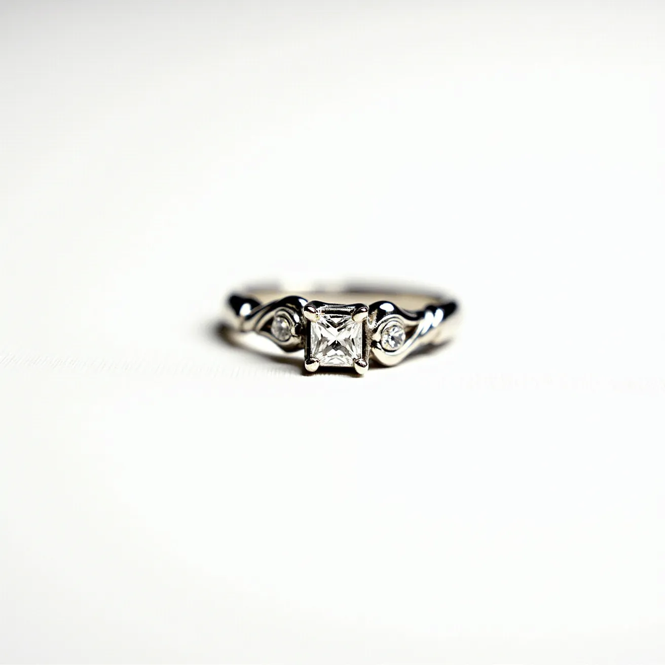 This silver wedding ring features a central princess-cut gemstone, likely a diamond, set prominently in a four-prong setting. Flanking the central stone are two smaller round gems on either side, also set securely within the band, contributing to an elegant, classic design. The band itself is polished and gently curves, enhancing the overall aesthetic with its graceful lines.