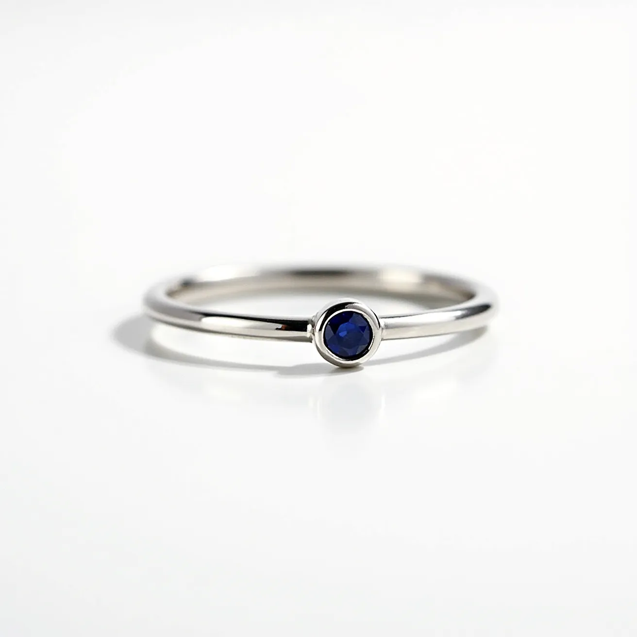 This solitaire ring features a sleek band crafted from what appears to be a reflective metal, likely white gold or platinum, known for its durability and lustrous shine. At its center, the ring showcases a single, vividly colored blue gemstone, potentially a sapphire, cut into a round shape. The stone is secured in a bezel setting, which encircles the gem with metal, providing protection and a modern aesthetic. The simple design emphasizes the solitary stone, allowing it to be the focal point without additional embellishments or clasps.