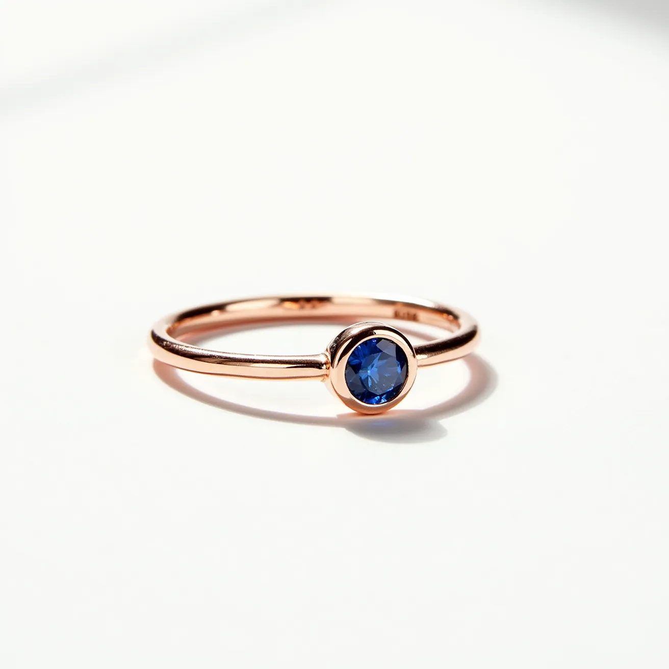 This solitaire ring features a band crafted from a rose gold material, giving it a warm and elegant appearance. The center of the ring showcases a striking blue sapphire, cut in a round brilliant style, which enhances its natural shine and reflective properties. The sapphire is securely set in a bezel setting, encircling the gemstone with a smooth metal rim that provides both protection and a modern aesthetic. This minimalist design allows the vibrant stone to be the focal point, creating a timeless and sophisticated piece of jewelry.