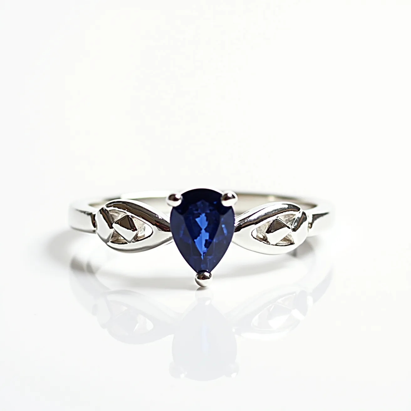 This solitaire ring features a beautifully crafted band made of a shiny, silver-colored metal. At its center is a pear-shaped blue gemstone, likely a sapphire, which is secured by a three-prong setting allowing the stone to be prominently displayed. The band is adorned on each side of the central gemstone with delicate metalwork resembling leaves or marquise shapes, enhancing the overall elegance of the design. The ring’s construction appears seamless with no visible clasps or attachments, emphasizing the minimalist and refined aesthetic.