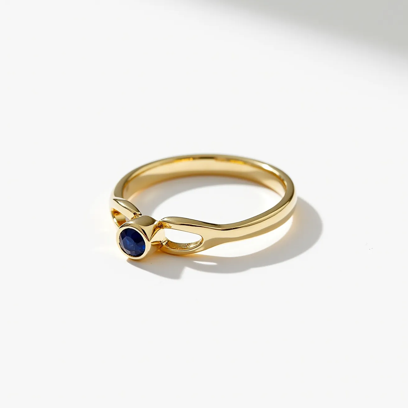 This solitaire ring features a sleek gold band with a modern, elegant design. The focal point of the ring is a deep blue round gemstone, likely a sapphire, securely held in place by a bezel setting that encircles the stone, enhancing its protection and elegance. The band itself has a smooth and polished finish, complementing the vibrant hue of the gemstone while adding a touch of timeless sophistication to the piece. The seamless design without any additional clasps or attachments emphasizes the simplicity and beauty of the solitaire setting, making it an exquisite piece of jewelry.