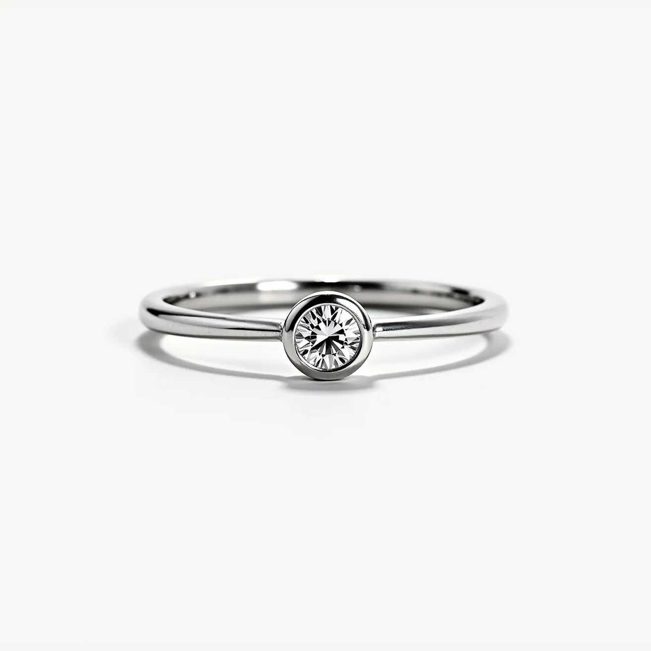 This solitaire ring is crafted from polished metal with a sleek, minimalist design. It features a single round cut diamond centrally positioned in a bezel setting, which encircles the gem with a protective band of metal while allowing ample light to enhance its brilliance. The band is smooth and slender, complementing the understated elegance of the ring. The seamless design emphasizes the diamond's clarity and sparkle, presenting a timeless piece suitable for various occasions.