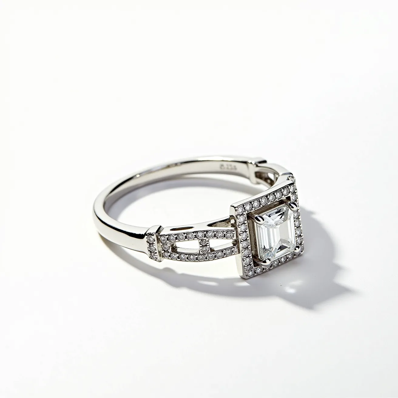 This square ring features a sleek metal band, likely crafted from a polished white gold or platinum, that enhances its elegance. At its center is an emerald-cut gemstone, possibly a diamond, securely set in a halo of smaller round-cut diamonds that accentuate the central gem. The square arrangement of the stones around the main gem adds a modern touch, complemented by additional small stones embedded along the band in an elaborate, openwork design. The combination of the cut, setting style, and arrangement of the stones displays a blend of classic and contemporary aesthetics.