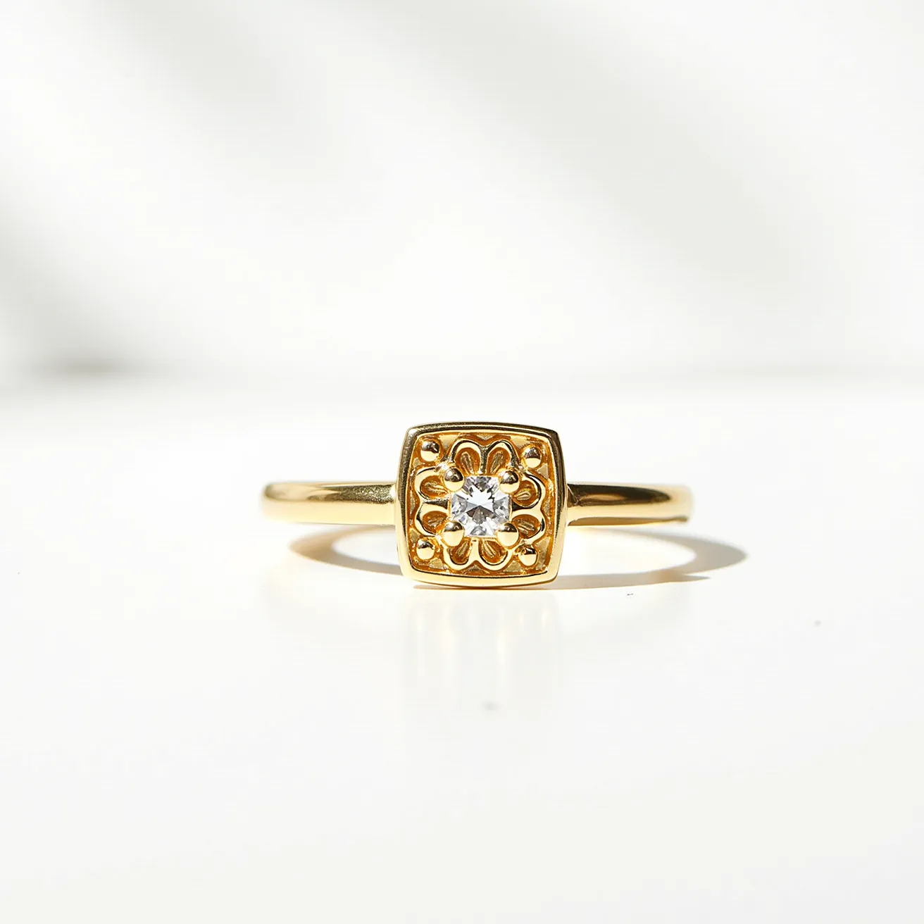 This square ring features a sleek gold band, highlighting a central square-shaped setting with a decorative floral design. At its center, a round-cut gemstone is securely nestled, providing a touch of elegance and sparkle. The stone is held in a bezel setting, which adds a contemporary touch to the overall design while ensuring durability. The ring displays a seamless integration of materials and design, offering both visual appeal and functional wearability.