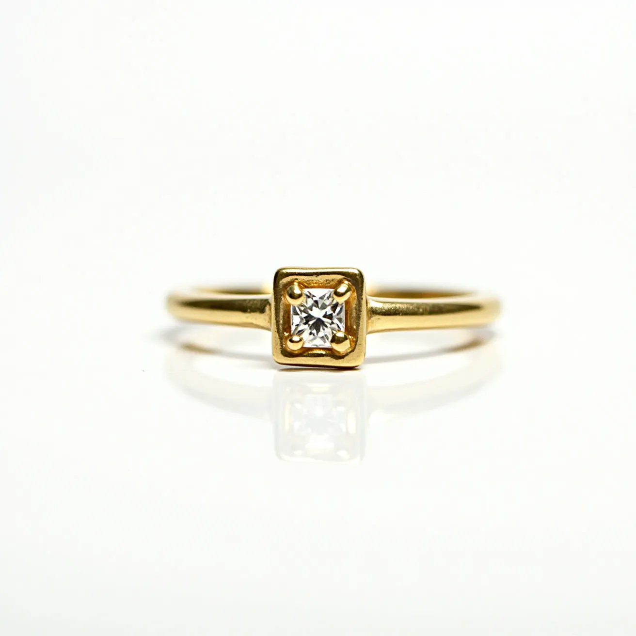 This square ring features a gold band with a minimalist design, showcasing a central square setting that holds a small, round-cut diamond. The setting is a bezel type, where the gold encircles the stone, providing a secure and elegant frame for the diamond. The smooth, polished gold adds a touch of sophistication, enhancing the overall aesthetic appeal. The ring's simplicity and fine craftsmanship make it a timeless piece, combining classic elegance with contemporary style.