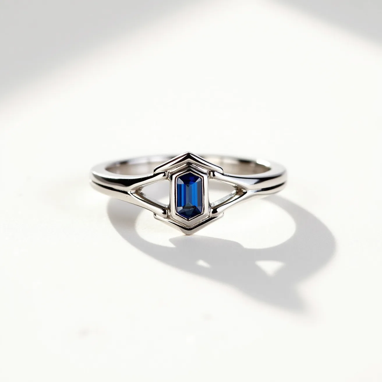 This square ring features a sleek and polished metal band, possibly made from silver or white gold, which complements its modern, geometric design. At its center lies a striking blue gemstone, likely a sapphire, cut into an elongated emerald shape and securely set within a bezel setting. The setting enhances the stone’s rich color and sophisticated appearance, offering both elegance and durability. The symmetrical design elements around the stone add a sense of balance and style to the piece, making it an eye-catching accessory.