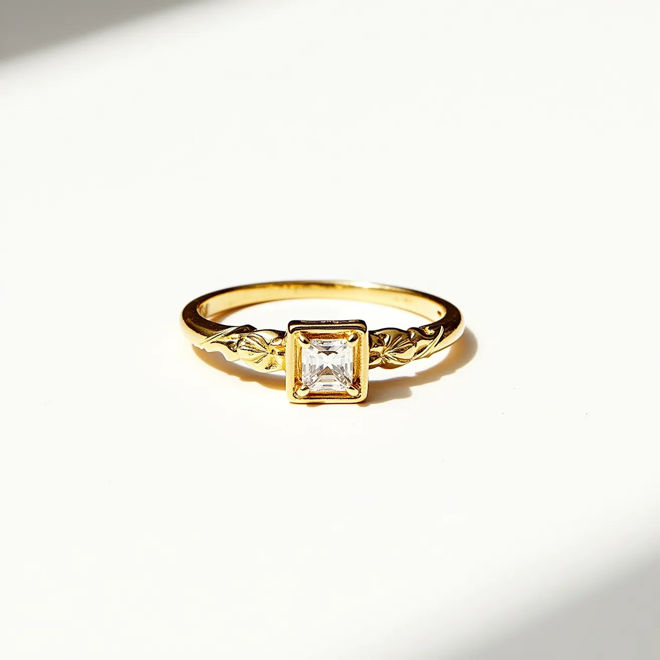 This square ring features a gold band with intricate leaf detailing on either side, highlighting its elegant and classic design. At the center, a square-cut gemstone, likely a diamond, is securely set in a bezel setting, adding a touch of sophistication and brilliance to the piece. The use of gold as the main material enhances the ring's luxurious feel, while the smooth finish of the band complements the sparkling central gem. The overall design merges traditional and contemporary elements, making it a versatile accessory for various occasions.