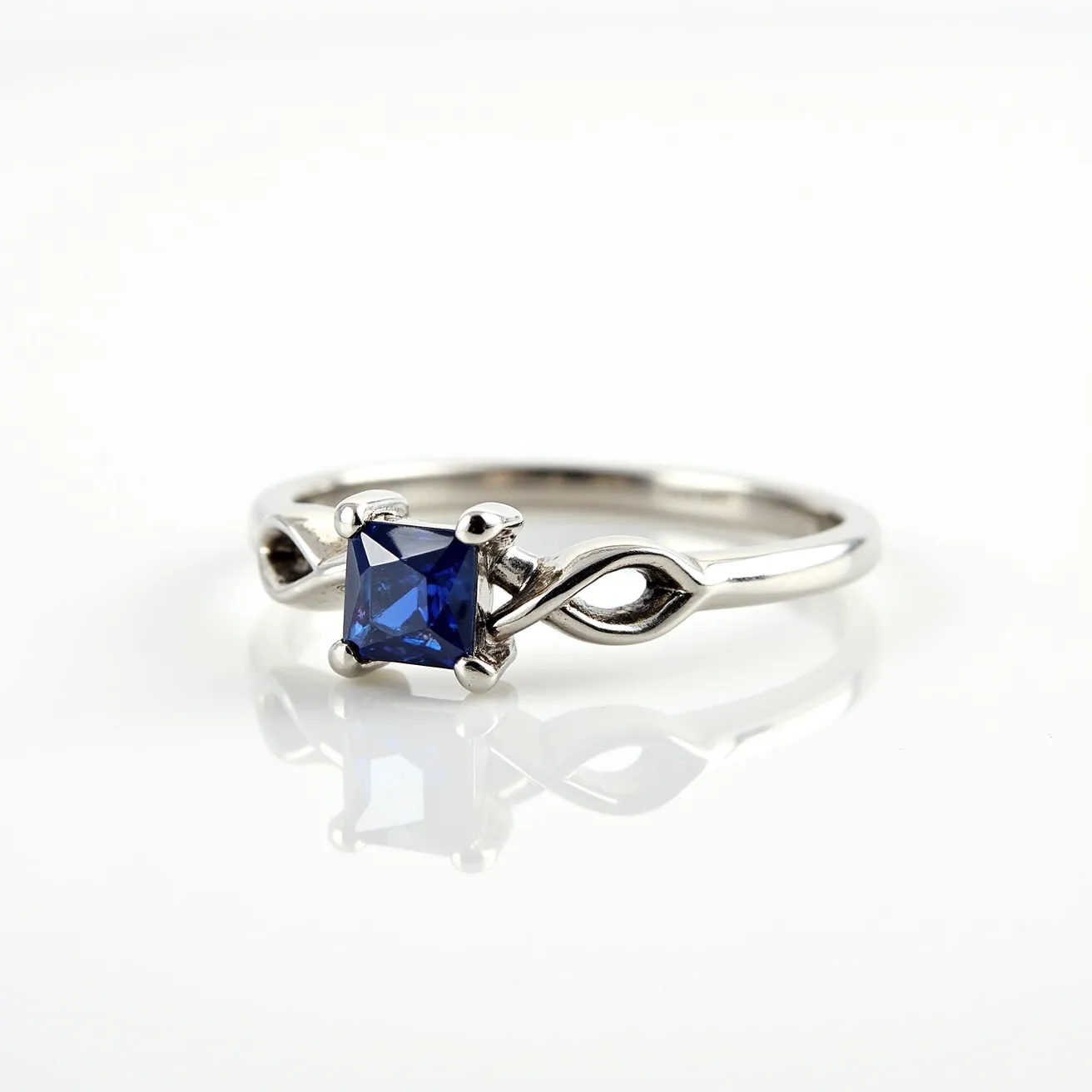 This square ring features a sleek, metallic band with a twisted design, most likely crafted from a silver-colored metal such as white gold or platinum. At its center, the ring showcases a square-cut blue sapphire, securely held in place by a four-prong setting that accentuates the gemstone’s deep color and precise cuts. The band’s interwoven design adds an elegant, flowing aesthetic, emphasizing both the gemstone and the craftsmanship of the metalwork. The ring lacks any additional clasps or attachments, maintaining a simple yet refined appearance.