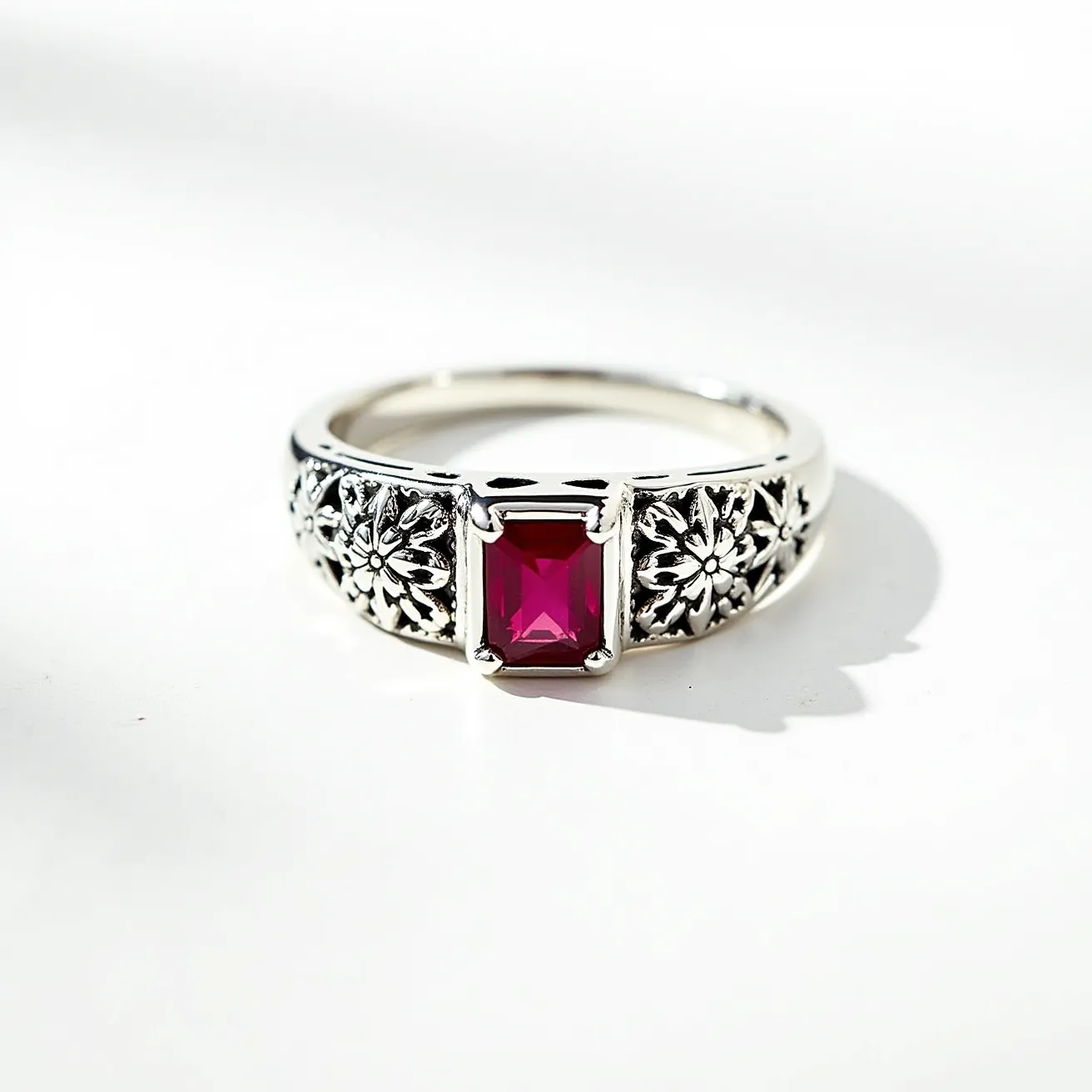 This square ring features a striking rectangular gemstone in the center, likely a ruby, exhibiting a rich red hue. The stone is cut in an emerald shape, positioned in a secure prong setting that enhances its elegance and prominence. The band is crafted from a shiny metal, possibly sterling silver, showcasing intricate floral engravings on either side of the central gem. These embellishments add a touch of sophistication and detail, blending vintage and contemporary aesthetics seamlessly. The ring does not appear to have any clasps or attachments, relying instead on its ornamental design to make a statement.