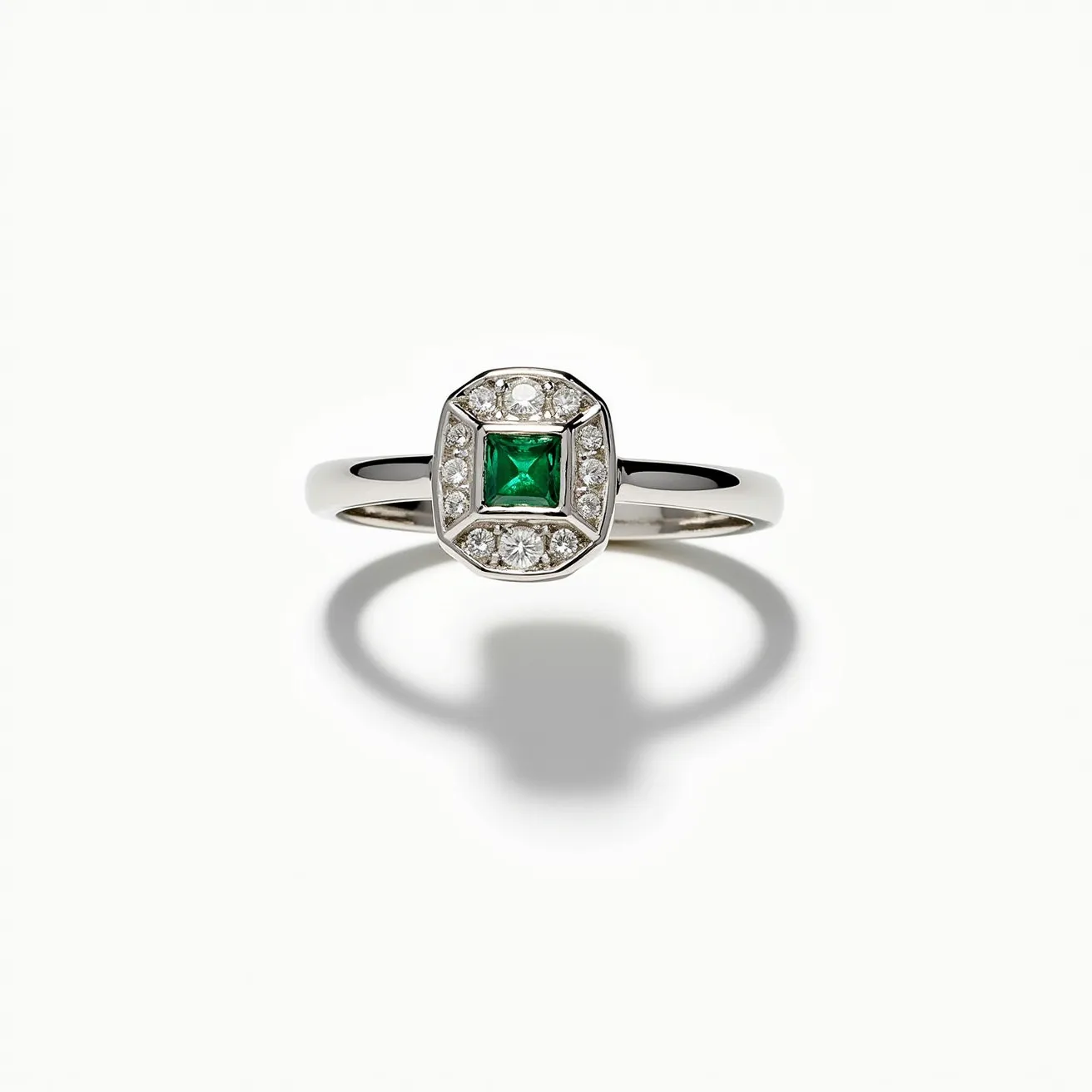 This square ring features a central, square-cut green gem, likely an emerald, set in the middle. Surrounding the central stone are multiple smaller, round-cut clear stones, presumably diamonds, arranged in a halo style. The ring appears to be made of polished metal, possibly white gold or platinum, which enhances the brilliance of the gemstones. The design is elegant and classic, with the stones securely held in place by a bezel-like setting, ensuring durability and stability. The band is smooth and reflective, completing the sophisticated look of the piece.