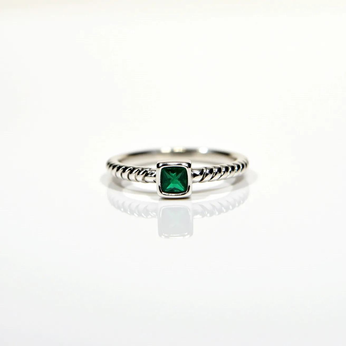 This square ring is crafted from a polished silver metal, featuring a central square-cut green gemstone, likely an emerald, set in a bezel setting. The band of the ring displays a twisted rope design, adding an element of texture and elegance. The stone's vibrant color provides a striking contrast against the sleek, reflective surface of the band, enhancing its overall visual appeal. The absence of a clasp or attachment suggests a traditional closed-loop construction, typical for rings of this style.