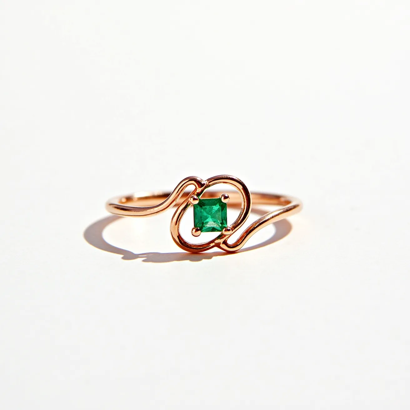 This square ring features a delicate and elegant design, crafted from a material that appears to be rose gold, indicated by its warm, pinkish hue. At its center, the ring displays a vibrant green square-cut gemstone, likely an emerald, which is set in a four-prong setting that securely holds the gem in place. The band of the ring exhibits a soft, flowing design that subtly enhances the overall aesthetic, wrapping gently around the central stone without any visible clasps or additional attachments. The meticulous craftsmanship highlights the stone's rich color, making it the focal point of this beautiful piece.