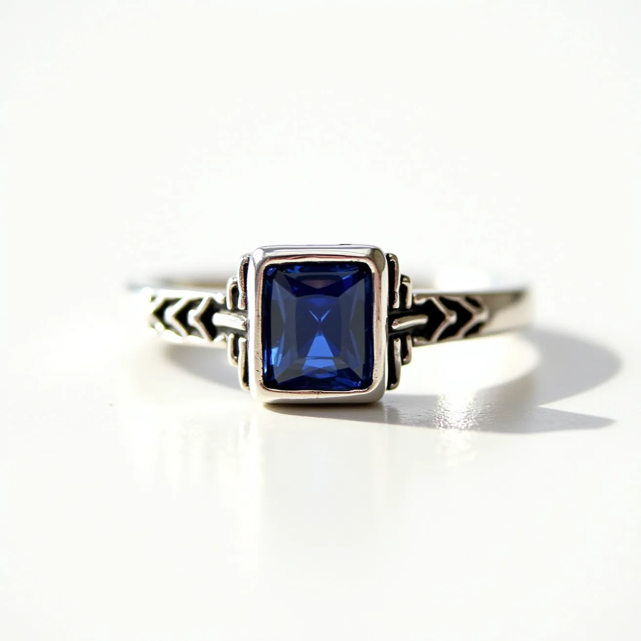 This square ring features a deep blue, princess-cut gemstone set prominently at its center. The gem is securely held in place by a bezel setting, which is framed by distinct, decorative metalwork on the band. Crafted from what appears to be a silver-toned metal, the band showcases intricate engravings resembling arrow patterns that add a unique character to the ring’s overall design. The thoughtful design and rich color of the gemstone create a striking visual appeal.