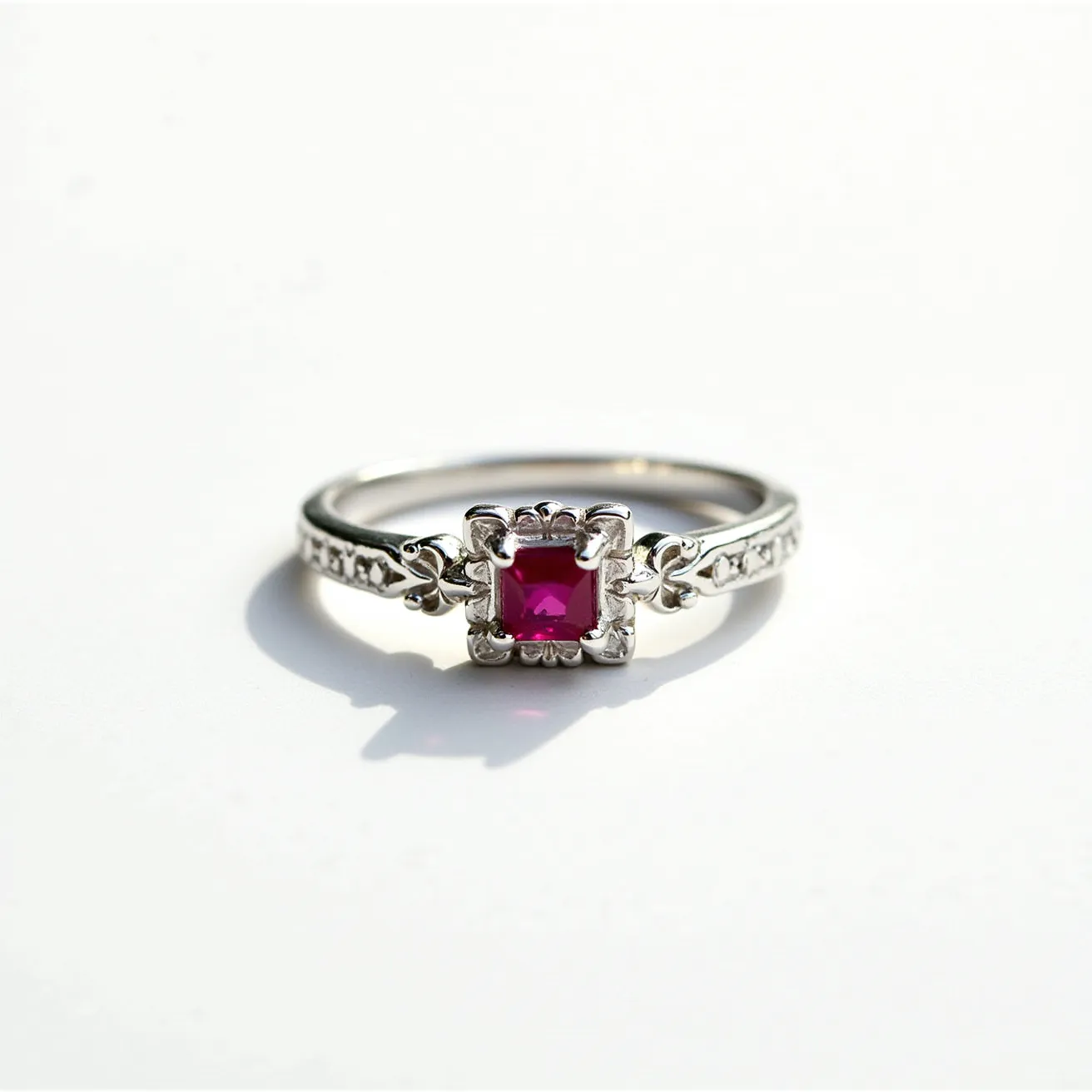 This square ring features a prominent red gemstone, likely a ruby, with a square cut set in an ornate silver setting. The gemstone is secured with detailed prongs that display intricate design work, complementing the overall elegance of the piece. The band is crafted from silver metal, embellished with fine detailing that adds to its sophistication and vintage charm. There is no visible clasp or attachment, emphasizing its simplicity and focus on the central gemstone.