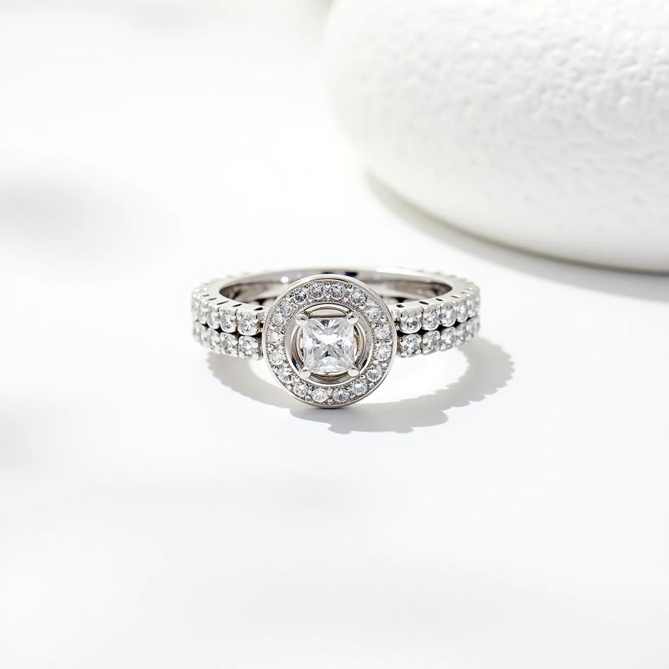 This stackable diamond ring features a central square-cut diamond encased within a halo of smaller round diamonds, creating a radiant centerpiece. The band is composed of metal, likely white gold or platinum, which complements the diamonds' brilliance. Adorning the band are two rows of pavé-set round diamonds that continue the sparkle around the finger. The overall design is elegantly cohesive, allowing for wear alone or stacked with other rings, with no visible clasps or attachments disrupting the seamless look.