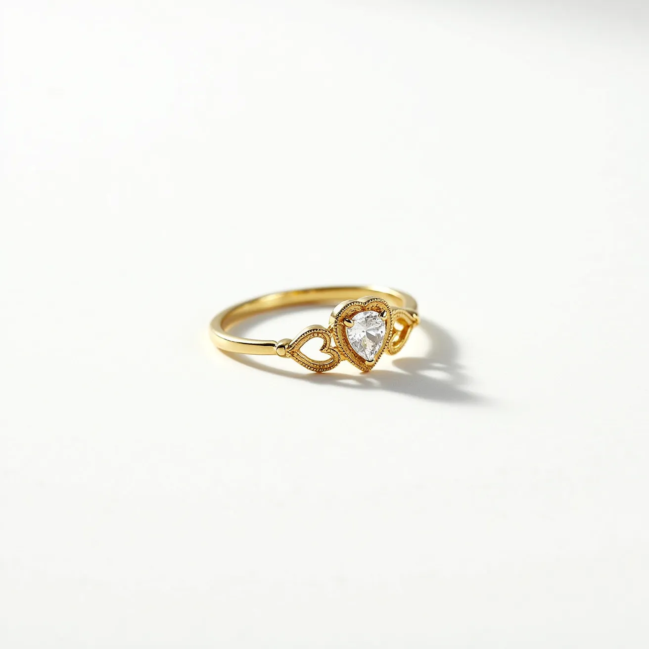 This stackable engagement ring features a delicate gold band, adorned with a heart-shaped gemstone as its centerpiece. The gemstone is set in a bezel setting, surrounded by intricate milgrain detailing that enhances its elegance. Flanking the center stone are two heart motifs crafted in the gold band, which complement the overall design and maintain the romantic theme of the piece.