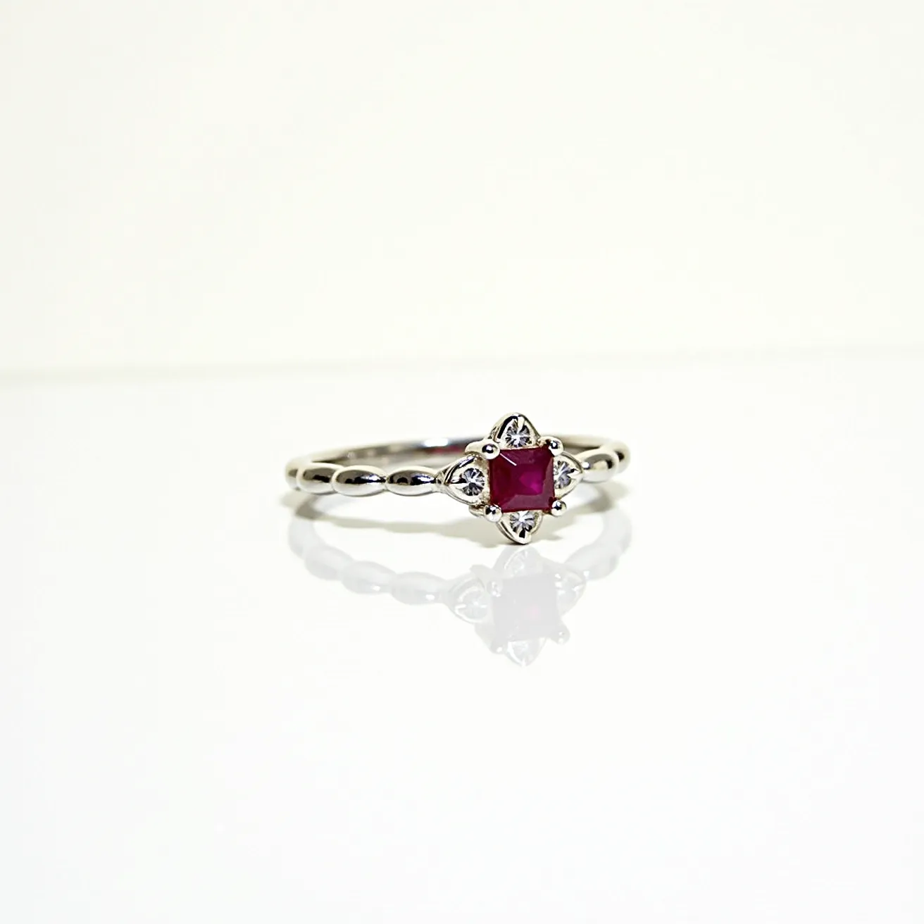 This stackable engagement ring features a delicate band crafted from a shiny metal, likely silver or white gold, which is formed into a series of smooth, rounded segments. At the center of the ring is a square-cut ruby, set in a bezel adorned with four small prongs that secure the gem in place. The ruby is surrounded by intricate metalwork resembling a floral pattern, suggesting a vintage or bohemian design. The overall craftsmanship indicates a focus on elegance and subtlety, making it suitable for stacking with other complementary rings.