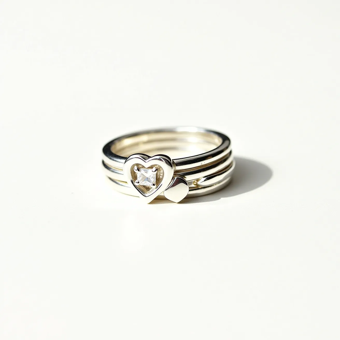 This stackable mothers ring features a sleek design composed of shiny silver bands. At the forefront is a prominent heart-shaped setting, which houses a clear, faceted stone. This stone is cut in a heart shape and securely set within the heart frame, adding a touch of elegance to the ring. Adjacent to the central heart is a smaller heart motif, also crafted in silver, complementing the overall theme. The rings are designed to nest together seamlessly, offering a versatile and sentimental piece of jewelry.