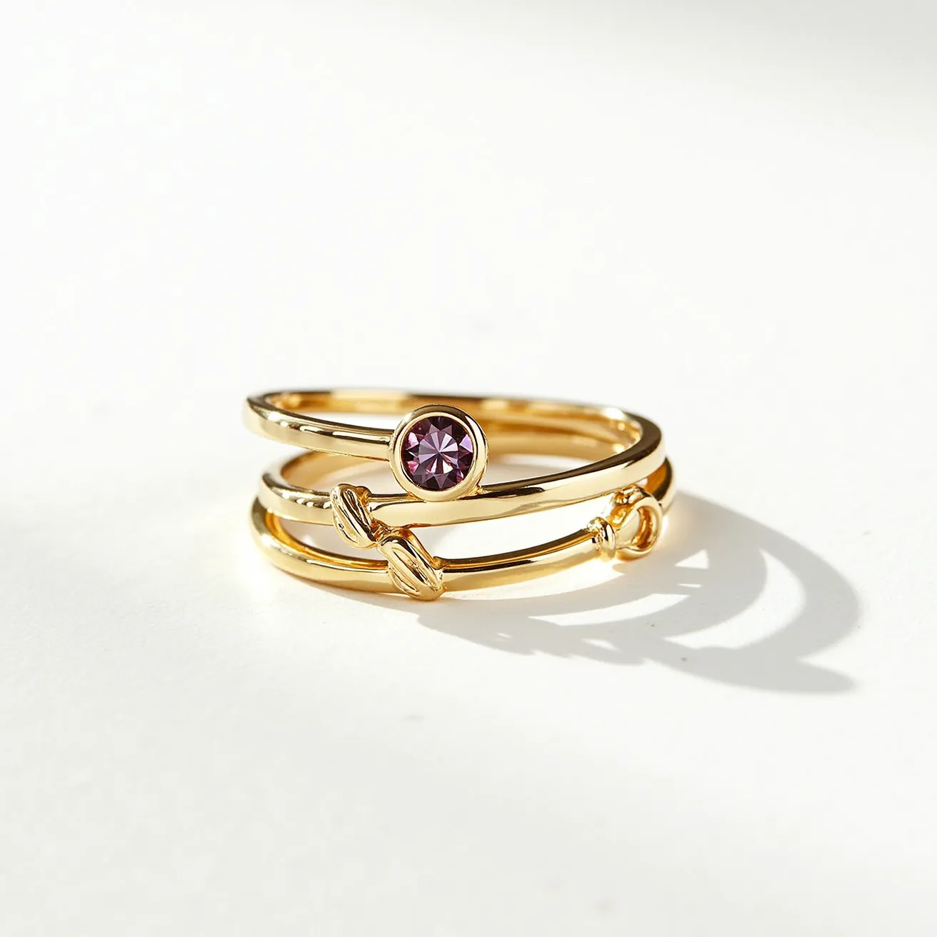 These stackable mothers rings are crafted from gold-toned metal, featuring a minimalistic and elegant design. The top ring showcases a round-cut purple gemstone securely set in a bezel setting, adding a touch of vibrant color. The adjacent ring is adorned with two decorative gold knots, providing additional visual interest and texture. The rings are designed to interlock seamlessly without the need for clasps or attachments, allowing them to stack neatly on the finger for a personalized and stylish look.