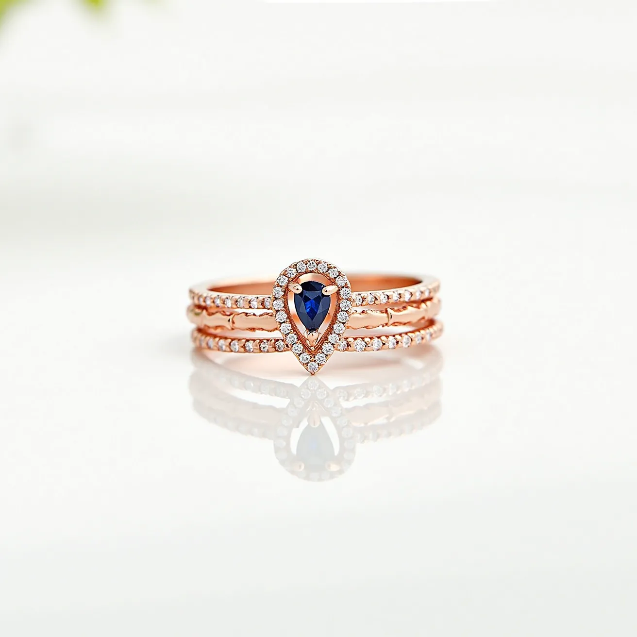 These stackable rings are crafted from a rose gold metal, featuring a central teardrop-shaped sapphire as the focal point. The sapphire is elegantly encircled by small round diamonds, forming a halo setting that enhances its brilliance. Three individual bands are stacked together, with each band adorned with additional small diamonds, creating a cohesive and sparkling design. The combination of the vibrant blue sapphire and the shimmering diamonds set against the warm rose gold offers a timeless and sophisticated look. There are no visible clasps or attachments as the rings are designed to slip onto the finger seamlessly.