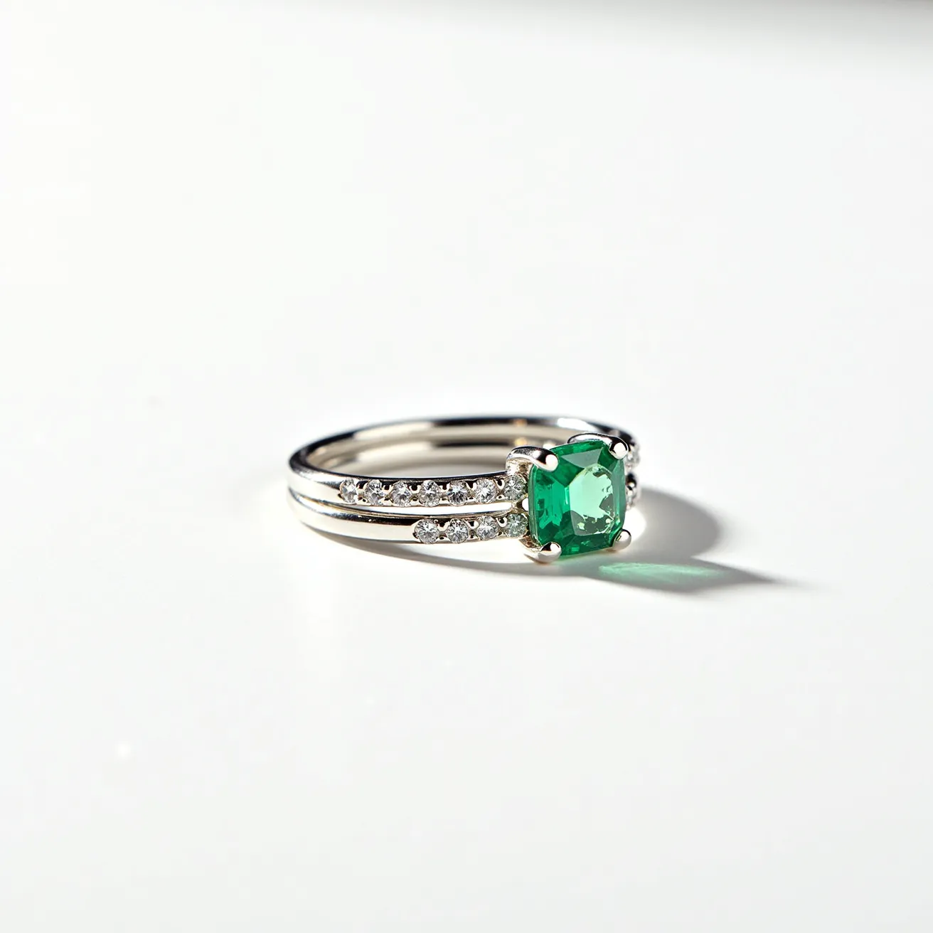 These stacked wedding rings feature a prominent square-cut green gemstone set in a claw setting, which beautifully occupies the center of attention. The metal bands appear to be crafted from a reflective material, likely a type of precious metal like white gold or platinum, providing a sleek, polished look. Enhancing the bands are small, round, clear stones, possibly diamonds, intricately set along the band in a pave setting, adding a touch of shimmering elegance. With a flawless blend of vibrant and subtle elements, these rings encapsulate a harmonious balance of color and brilliance.