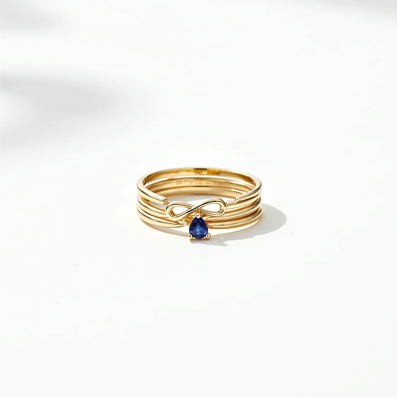 These stacked wedding rings are crafted from a polished gold material, featuring a harmonious set of slender bands. The focal point is a prominently set blue pear-shaped gemstone, likely a sapphire, secured in a prong setting that elegantly anchors it in place. An infinity symbol crafted from the same gold material is intertwined with the gemstone, adding a meaningful and decorative element to the composition. The rings are seamlessly stacked, with no visible clasps or attachments interrupting their sleek and unified appearance.