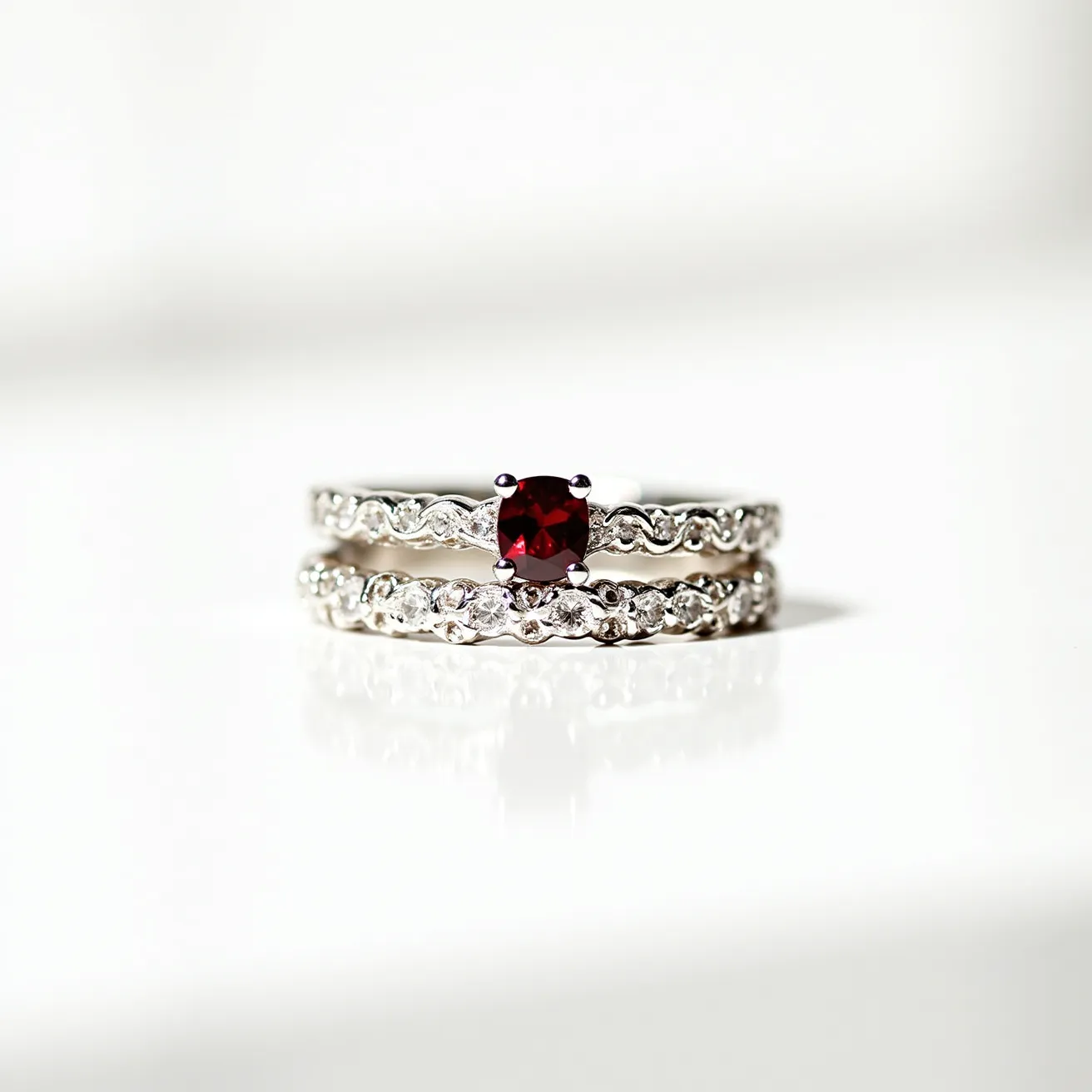 These stacked wedding rings feature intricately designed bands crafted from white gold. The top ring is adorned with a prominent round-cut red gemstone set in a classic four-prong setting, adding a vibrant focal point. Both rings are embellished with a series of small, round-cut diamonds that are set closely together along the band, creating a continuous line of sparkle that enhances their elegant appeal. The arrangement and setting of the gemstones suggest a meticulous attention to detail, complementing the overall sophistication of the rings.