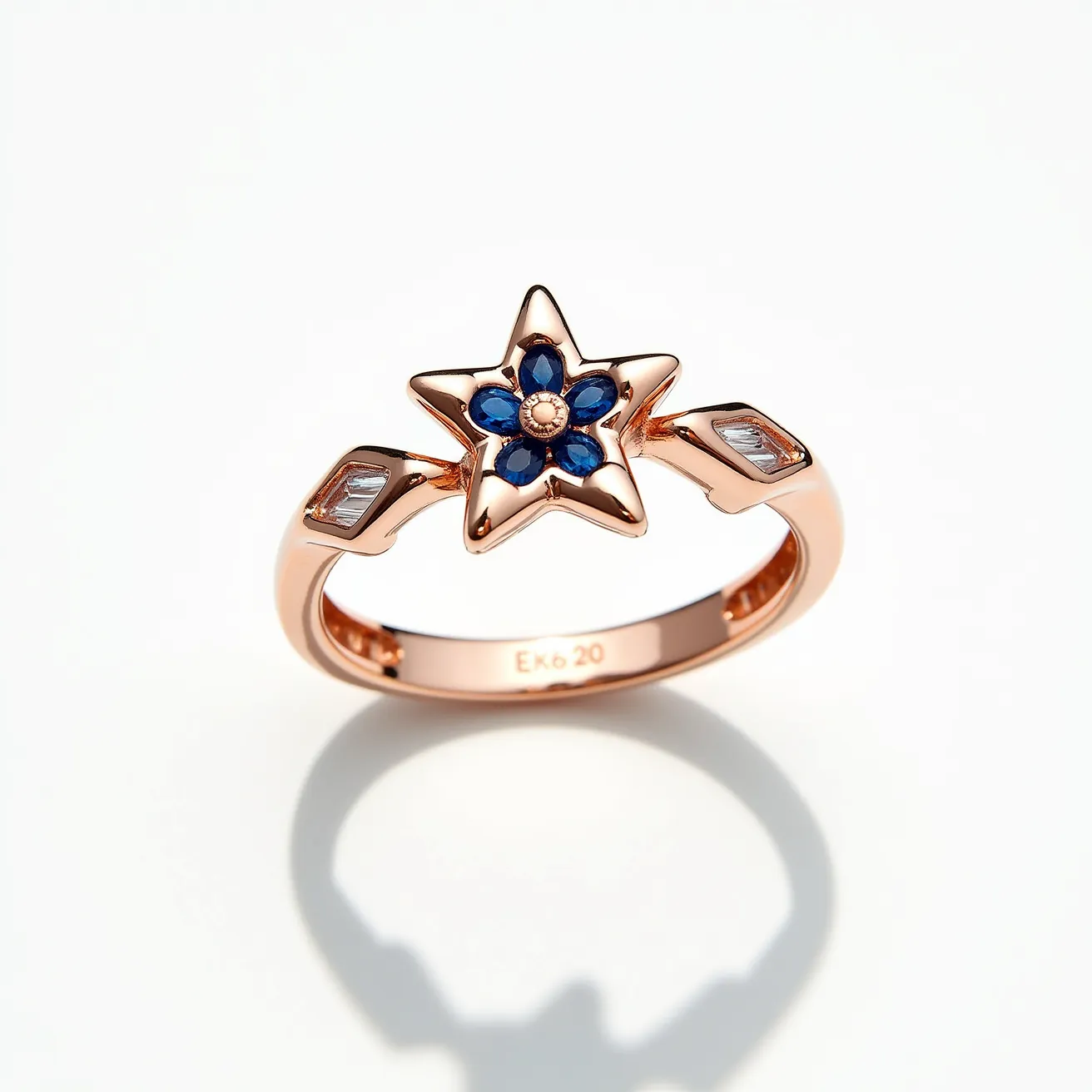 This star ring features a beautiful design crafted from rose gold, forming a striking star shape as its centerpiece. The star is adorned with blue gemstones, likely sapphires, arranged in a floral pattern with a small central gem creating a captivating focus. The gemstones appear to be round cuts, securely set within the star. Additionally, the band's shoulders are embellished with rectangular clear stones, possibly diamonds, set in a channel setting, adding an elegant touch to the overall design. The combination of materials and gems gives the ring a modern yet timeless appeal.