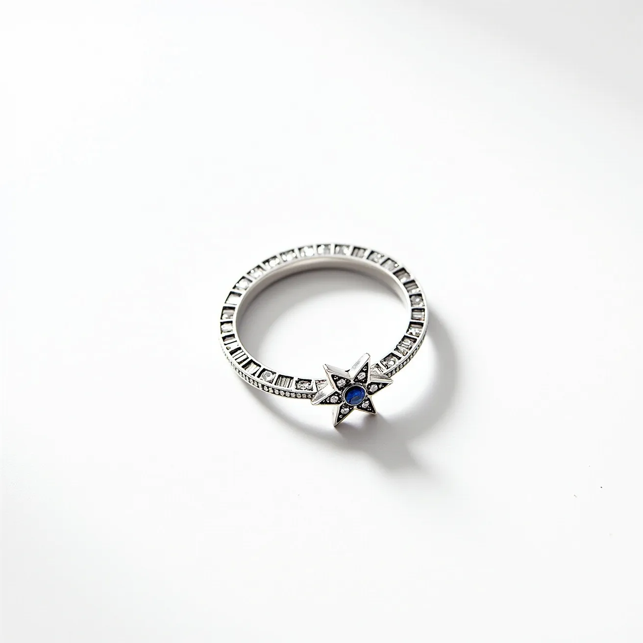 This star ring features a delicate and elegant design crafted from a polished silver or white gold material. At its focal point is a star-shaped setting adorned with a central round-cut blue sapphire, providing a captivating contrast against the metallic base. The star is further embellished with smaller, clear stones, likely diamonds or cubic zirconia, which accentuate the sparkle and allure of the ring. The band is skillfully designed with a series of square-cut stones, channel-set around the circumference, adding to its sophistication. The ring's seamless design suggests there is no visible clasp or attachment, maintaining a smooth and continuous aesthetic.