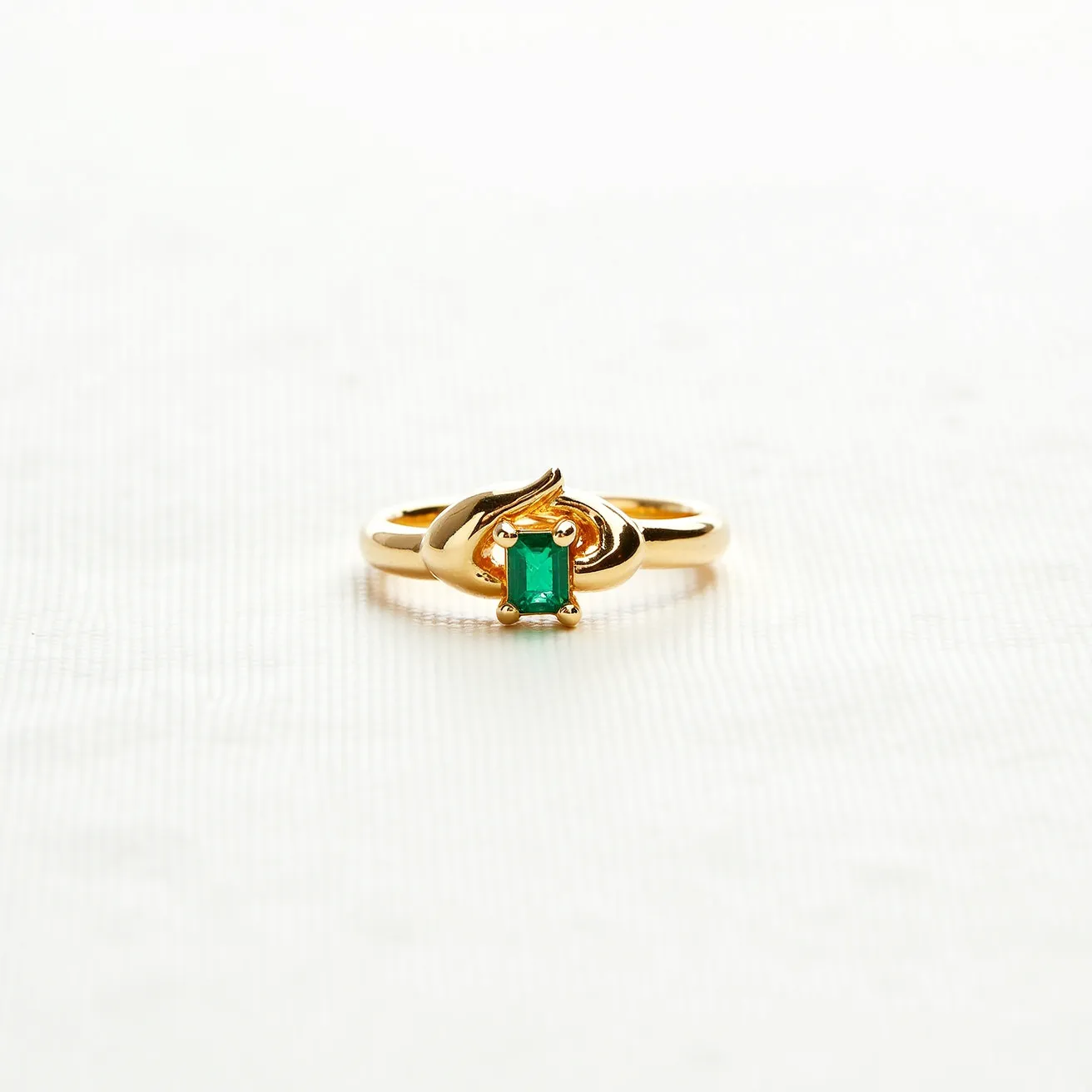 This statement ring features a refined and elegant design crafted from polished gold. At its center, it showcases an emerald-cut green gemstone, securely held in place by a four-prong setting that highlights the stone's vivid color and clarity. The band itself is smooth and subtly twisted, adding a dynamic and modern touch to the overall aesthetic. The simplistic yet striking design emphasizes the beauty of the gemstone, making it a timeless piece suited for any occasion.