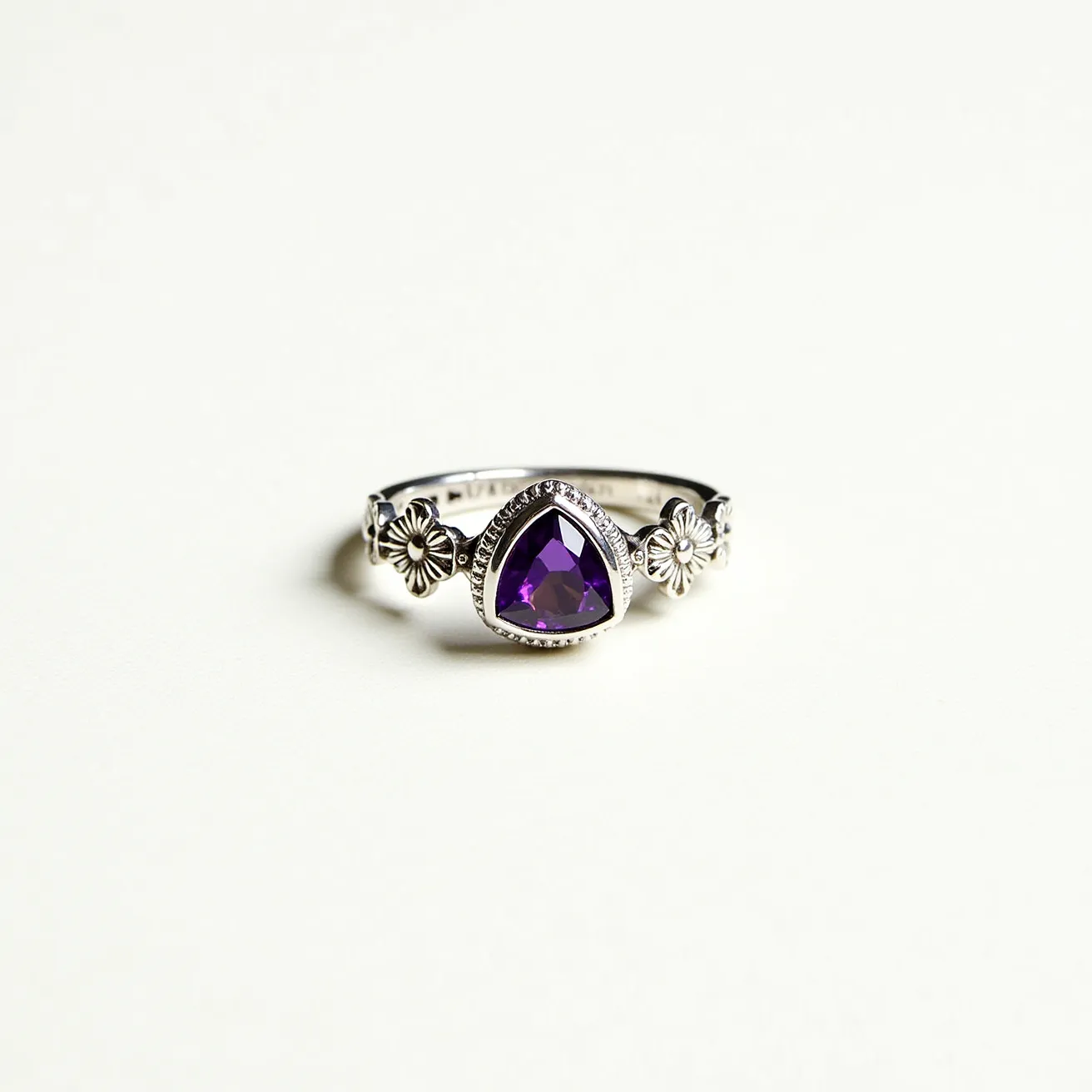 This statement ring features a striking design, prominently showcasing a triangular-cut amethyst stone at its center. The gemstone is set in a bezel setting, which highlights its deep purple hue and adds to its elegance. Flanking the amethyst are two intricately designed floral motifs, providing a vintage-inspired charm. The band appears to be crafted from a polished silver metal, which complements the overall aesthetic and enhances the brilliance of the amethyst. The combination of the gemstone with the floral accents creates a harmonious balance between boldness and delicacy.