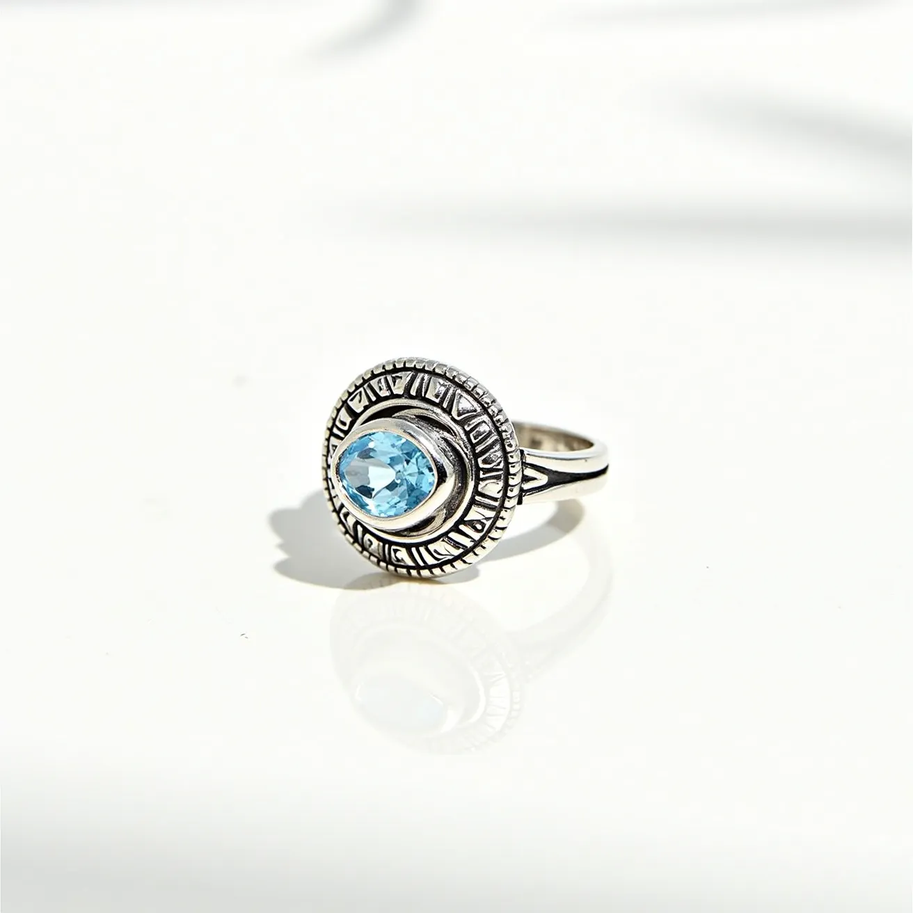 This statement ring showcases a striking blue gemstone, likely topaz, designed in an oval cut and set prominently at the center. The stone is embraced by an ornate bezel setting that adds an additional layer of intricate embellishment. The ring is crafted from what appears to be silver, featuring an engraved band with detailed motifs that complement the central stone. The craftsmanship highlights a blend of both modern and classical design elements, giving the piece a timeless appeal.