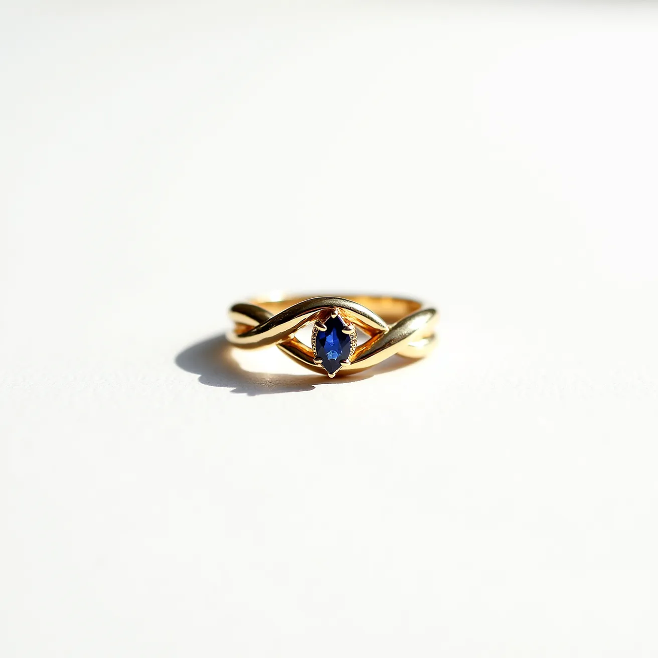 This tangled ring features an intricate gold band that twists elegantly, showcasing a central marquise-cut blue gemstone, likely a sapphire. The stone is securely held in place by a bezel setting, which emphasizes the gem's deep color and elongated shape. The gold band interweaves around the stone, adding dimension and depth to the design, while maintaining a classic and sophisticated appearance.