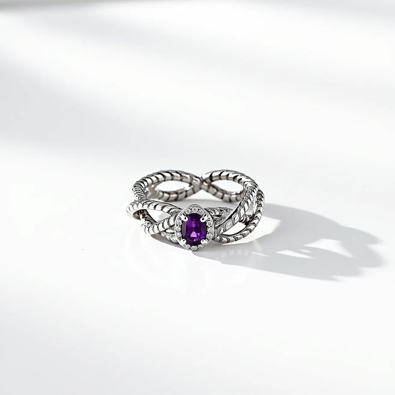 This tangled ring features an intricate design crafted from silver, showcasing a twisted rope motif that intertwines elegantly. At the center, a prominent oval-cut purple gemstone, likely an amethyst, is securely set in a bezel setting, surrounded by a halo of small, clear stones that are likely diamonds or cubic zirconia. The combination of the striking purple gem and the delicate accent stones adds a touch of elegance and sophistication to the piece. The ring's construction lacks a traditional clasp, designed as a continuous band for ease of wear.