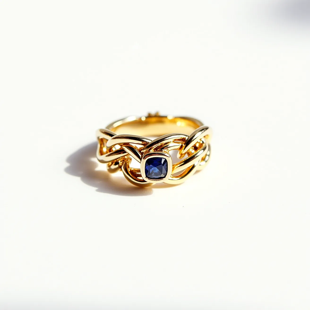 This tangled ring features an intricate design crafted from polished gold, characterized by its intertwined, looped bands. At the center, a striking deep blue sapphire is prominently displayed, cut in a cushion shape. The gemstone is set securely in a bezel setting, allowing the gold to encircle the sapphire with a smooth, protective frame. The ring does not appear to have any clasps or additional attachments, maintaining a seamless, continuous form.