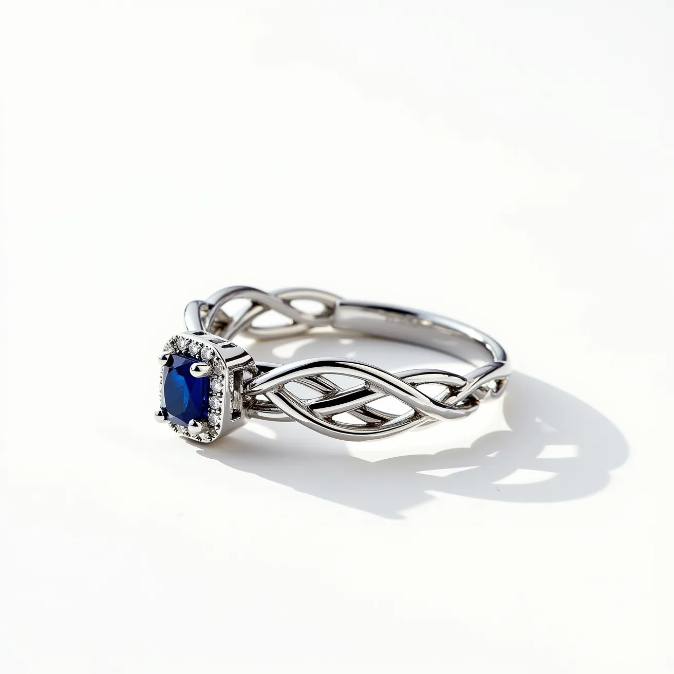 This tangled ring features an elegant, intricate design formed from a polished metal, likely white gold or platinum, that weaves together in a graceful pattern. At its center, there is a deep blue, cushion-cut stone, possibly a sapphire, surrounded by a halo of small, clear round-cut gems, likely diamonds, set in a prong setting that accentuates the central gemstone's color and cut. The ring's shank showcases a twisted, open-work design that adds to its delicate appearance, allowing light to play off the metal and stones beautifully. There is no visible clasp or attachment, suggesting it is a traditional ring crafted for aesthetic allure and wearer comfort.