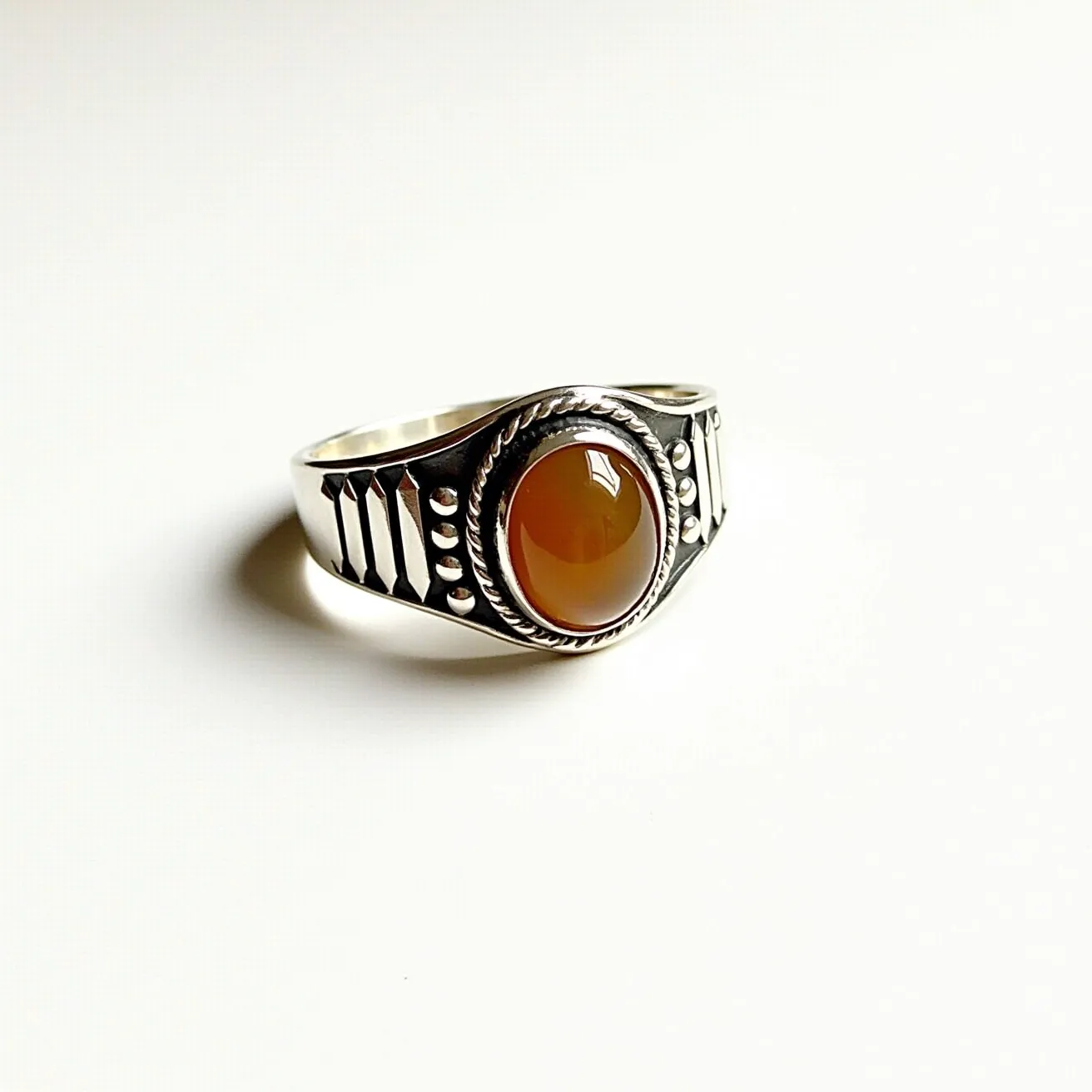 This thumb ring features a polished silver band adorned with decorative geometric engravings on the sides. At its center is a smooth, oval-shaped cabochon made of a warm, amber-colored stone, securely set into the band with a decorative twisted rope-style bezel setting. The design is both bold and elegant, showcasing a blend of traditional and modern aesthetics.