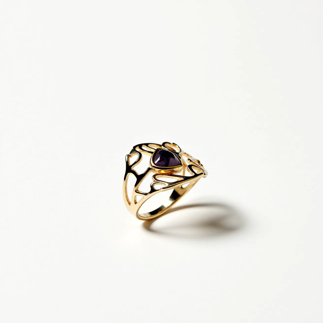 This thumb ring features an intricate design crafted from gold, showcasing an openwork pattern with heart motifs. At its center, a dark purple gemstone is set, likely amethyst, cut in a faceted shape to enhance its brilliance. The stone is embedded in a bezel setting which provides security and complements the overall aesthetic. The design is continuous and does not include any clasps or attachments, allowing for a smooth and elegant appearance.