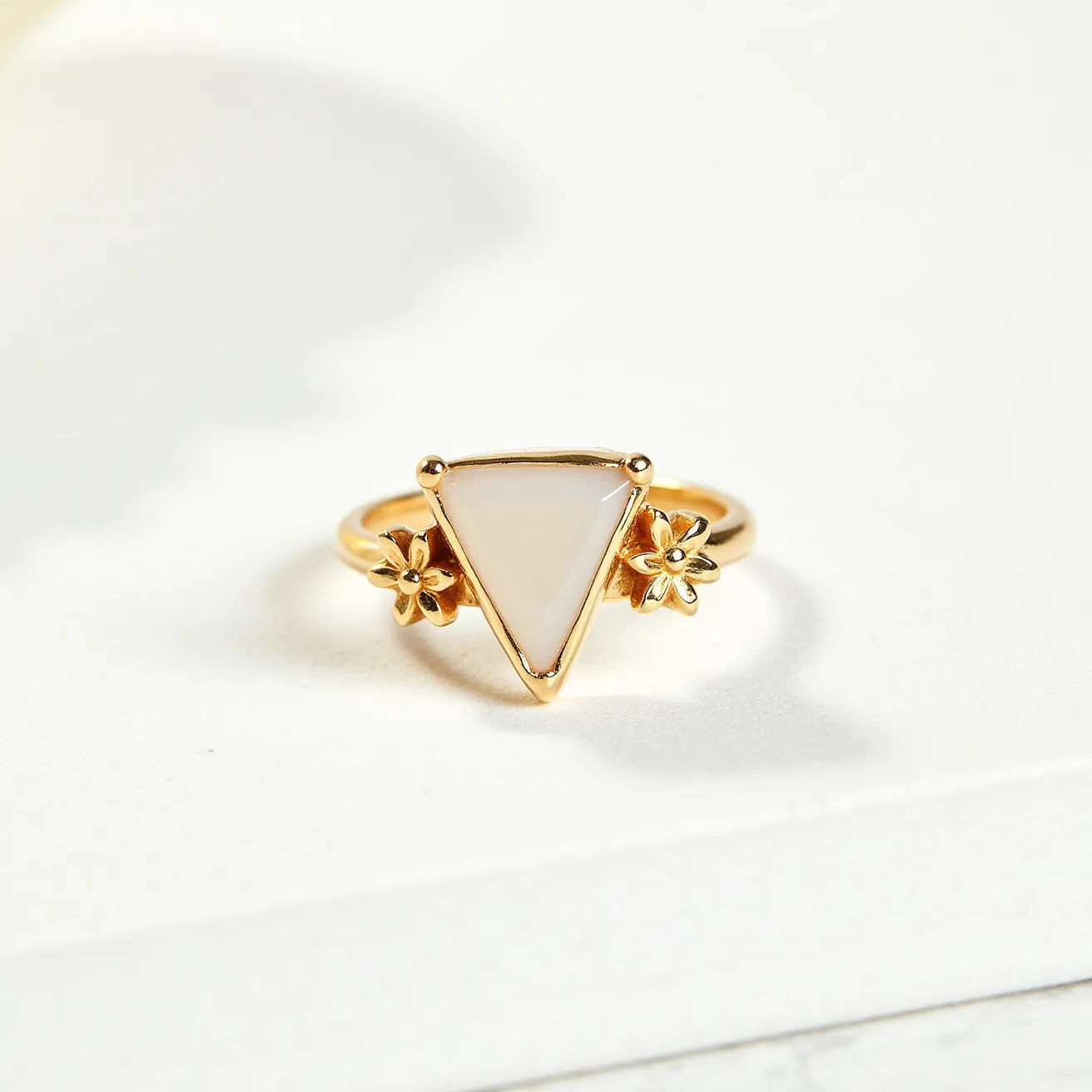 This thumb ring features a triangular gem in a bezel setting, crafted from a soft white material, perhaps a type of polished stone or glass. The gem is secured within a gold-toned metal band that adds a warm contrast to the gem. On either side of the central gem, there are delicate floral embellishments, also fashioned from the same gold-toned material, enhancing the design with a touch of whimsy and elegance. The ring appears to have a solid, continuous band without any clasps or additional attachments, emphasizing simplicity and style.