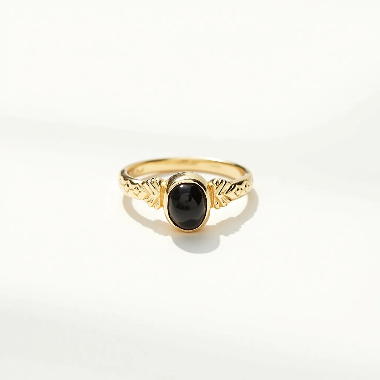 This thumb ring features a band made of polished gold with intricate leaf-like engravings on the shoulders, enhancing its elegant design. At the center sits a black oval cabochon gemstone securely set in a bezel setting, providing a smooth and seamless appearance. The combination of the dark stone with the gold band creates a striking contrast, adding a touch of sophistication to the piece. There is no visible clasp or attachment, indicating a standard ring design for easy wear.