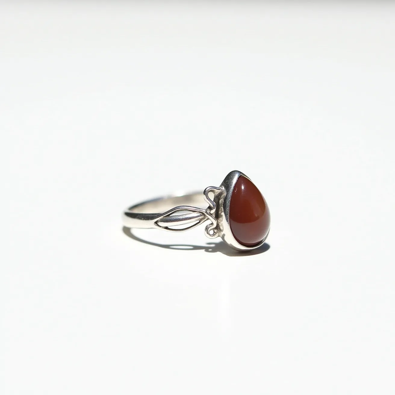 This thumb ring features a polished silver band with an intricate leaf design on either side of a teardrop-shaped stone. The gem appears to be a polished cabochon-cut carnelian, set securely in a bezel setting that enhances its warm, reddish-brown hue. The overall design is elegant and earthy, with the leaf motif adding a natural element to the piece. There is no clasp or additional attachment, emphasizing the simplicity and elegance of the ring’s design.