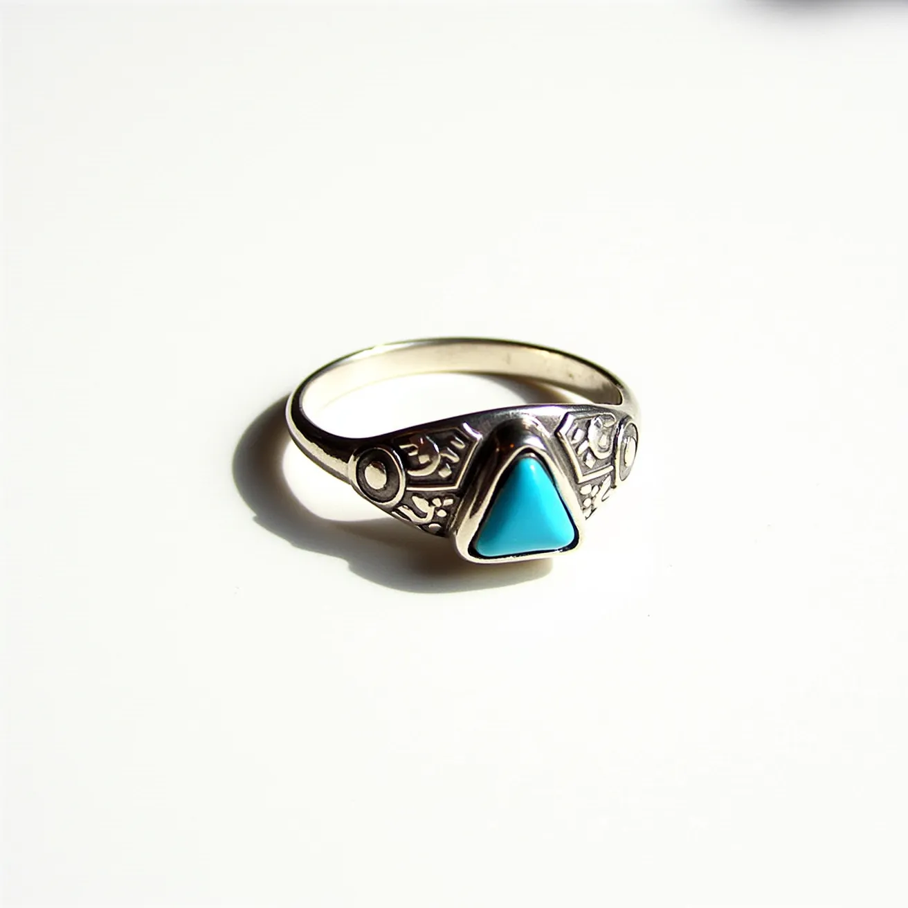 This thumb ring features a prominent triangular turquoise gemstone set in a bezel setting, with intricate engravings adorning the metal band on either side. The metal appears to be silver, enhancing the vibrant blue of the gemstone. The band has a smooth, polished finish, providing a contrast to the detailed engravings. The overall design is simple yet elegant, with the gemstone as the focal point, and the engravings add an element of artistry to the piece.